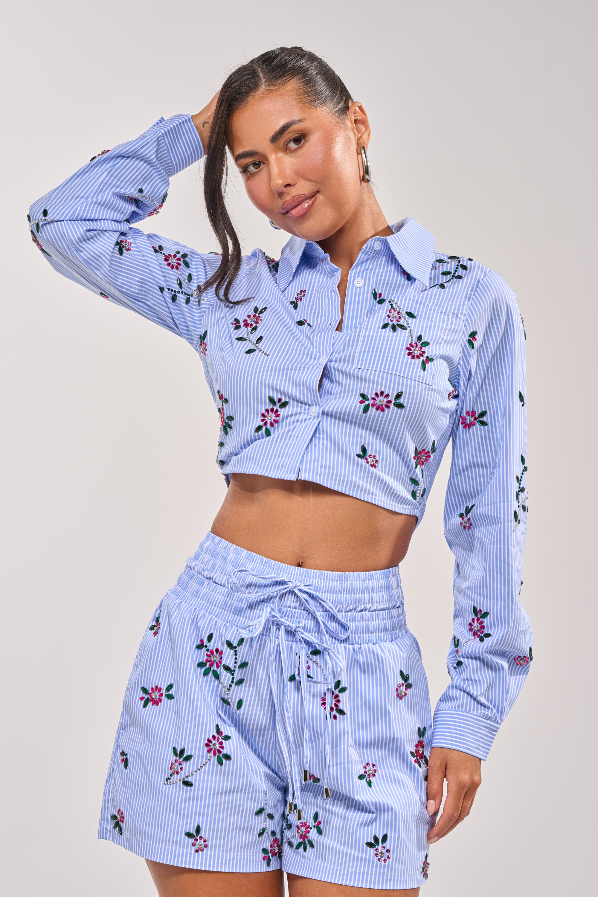 A woman models the JUNIE POPLIN RHINESTONE DETAIL BLOUSE, a blue striped, long-sleeve, collared crop top with rhinestone accents, paired with matching high-waisted shorts against a plain light background. 0