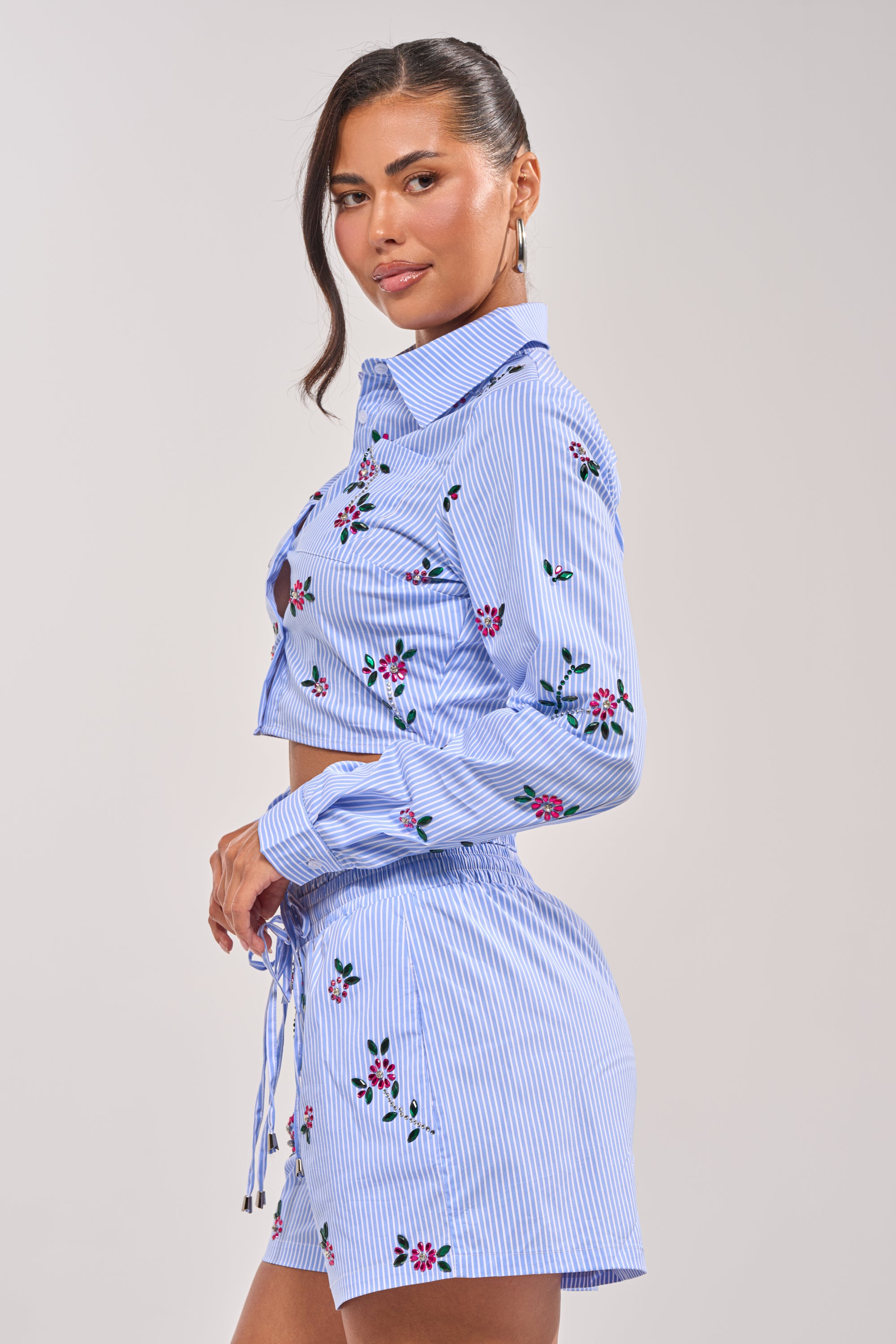 Woman standing sideways, wearing the JUNIE POPLIN RHINESTONE DETAIL BLOUSE—blue with white stripes, long sleeves, cropped fit, and rhinestone details—paired with matching high-waisted shorts; neutral background. 1