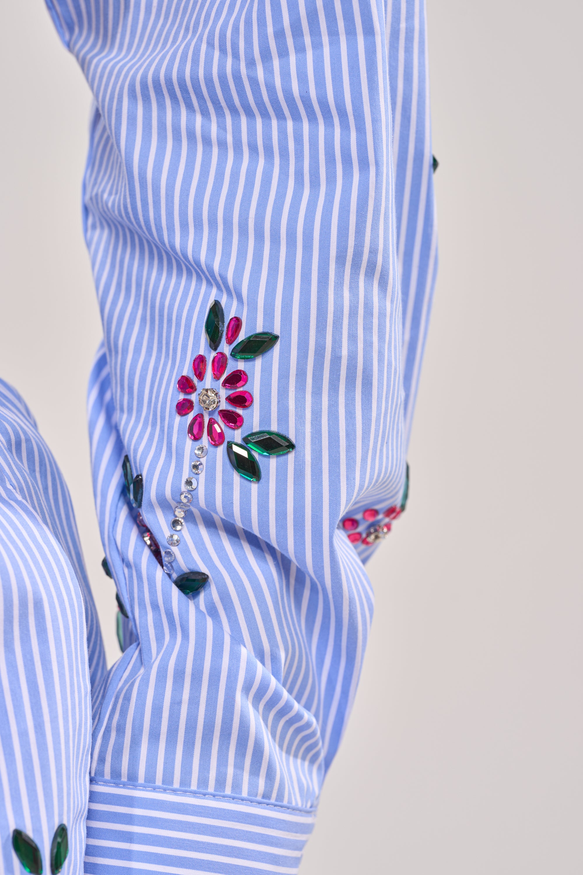A close-up of the JUNIE POPLIN RHINESTONE DETAIL BLOUSE sleeve shows blue stripes with pink, green, and silver embroidered and beaded floral accents—perfect for adding flair to any cropped button-down look. 5