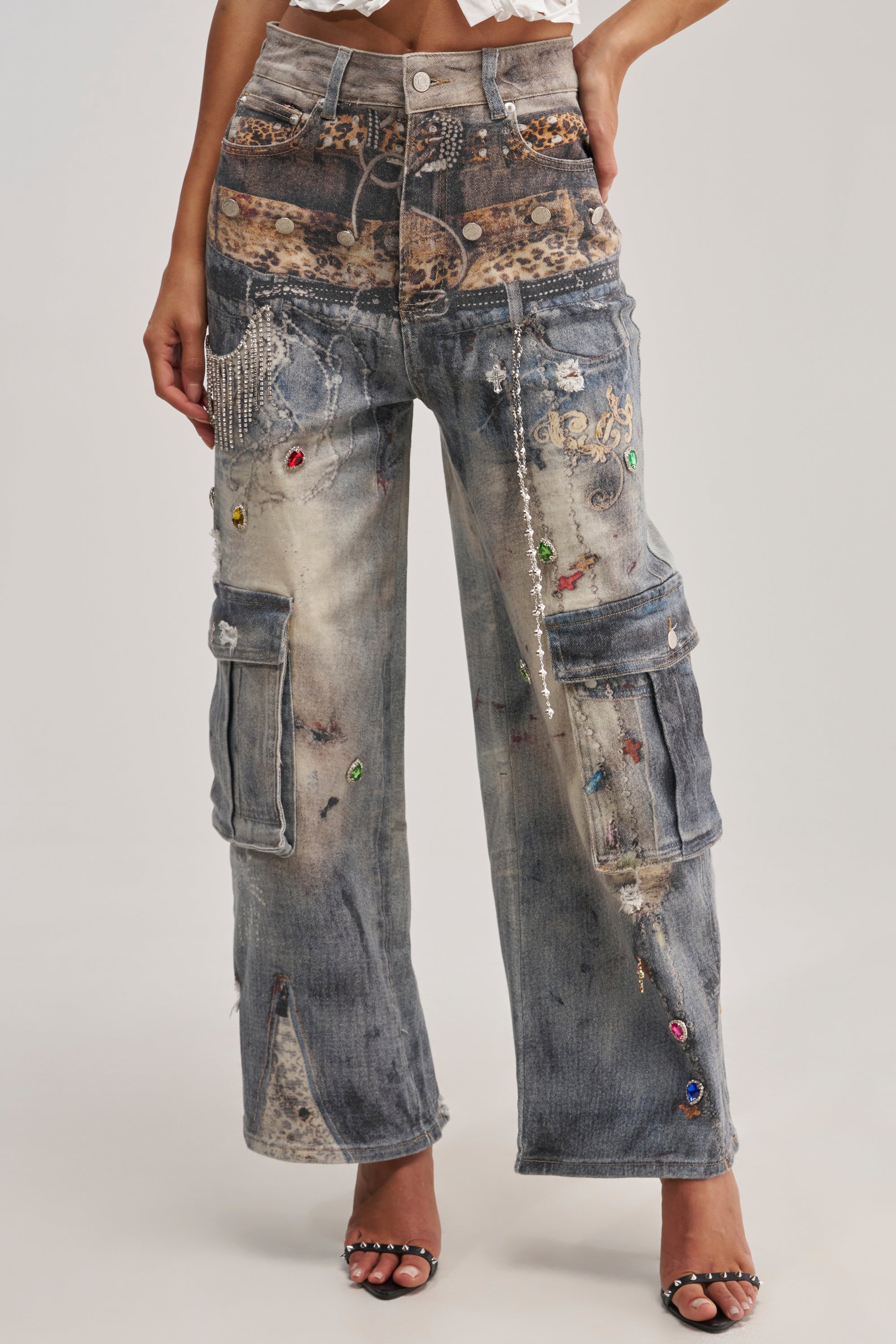 A person wears the COME AND GET IT RELAXED DENIM—distressed, loose-fitting maximalist jeans with embellishments, cargo pockets, colorful gems, lace patches, and chains—paired boldly with black heeled sandals.