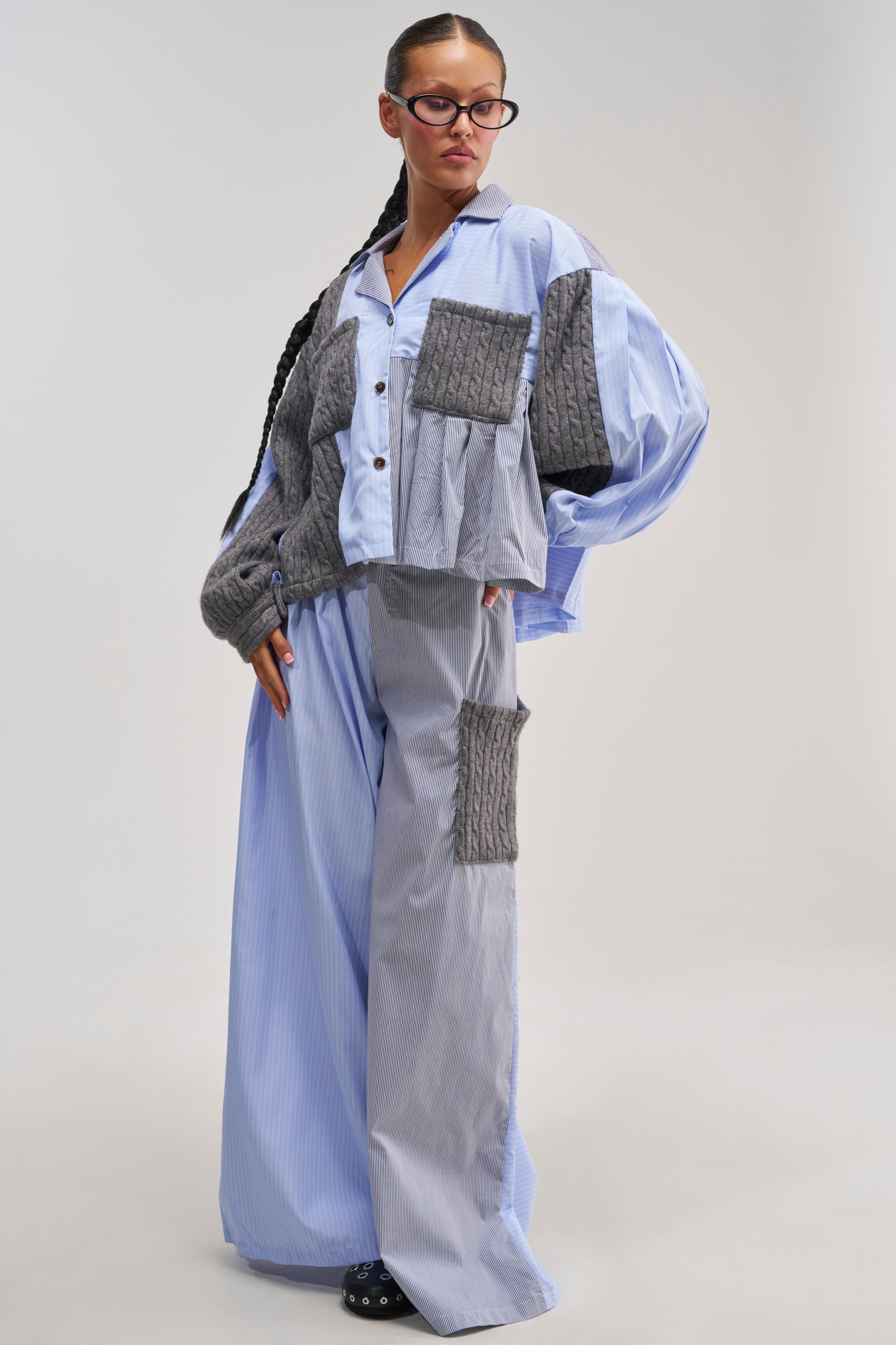 Wearing the SUNGGLES FOR EVERYONE TROUSER styled with an oversized blue pinstripe shirt in striped cotton poplin, black glasses, and dark shoes, this individual stands confidently against a plain light background—an effortlessly fashion-forward loungewear look.