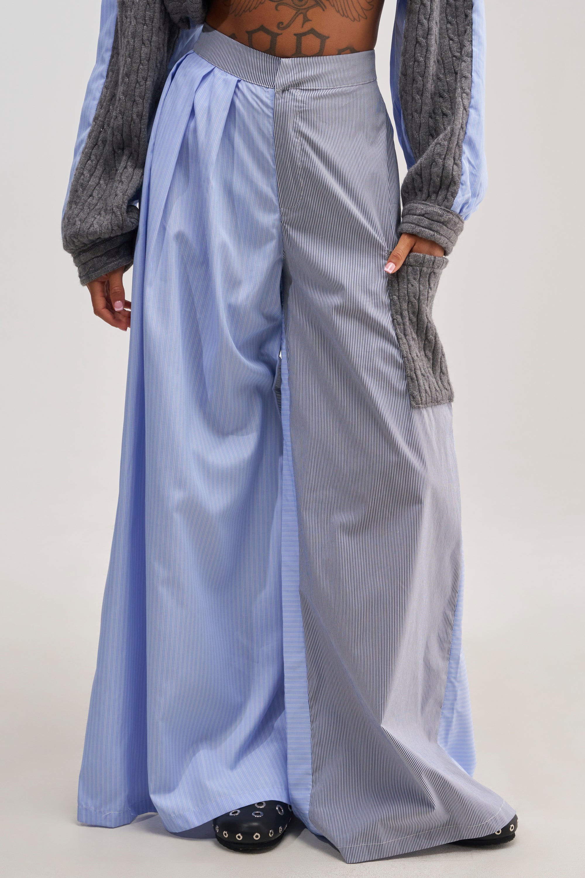 Person wearing the SNUGGLES FOR EVERYONE TROUSER—colorblock, wide-leg, high-waisted pants in blue and gray pinstripe, crafted from striped cotton poplin with a pleated design and mixed fabric panels for a fashion-forward loungewear vibe. The look is styled with long gray sleeves, a visible tattoo on the midriff, and black studded shoes.