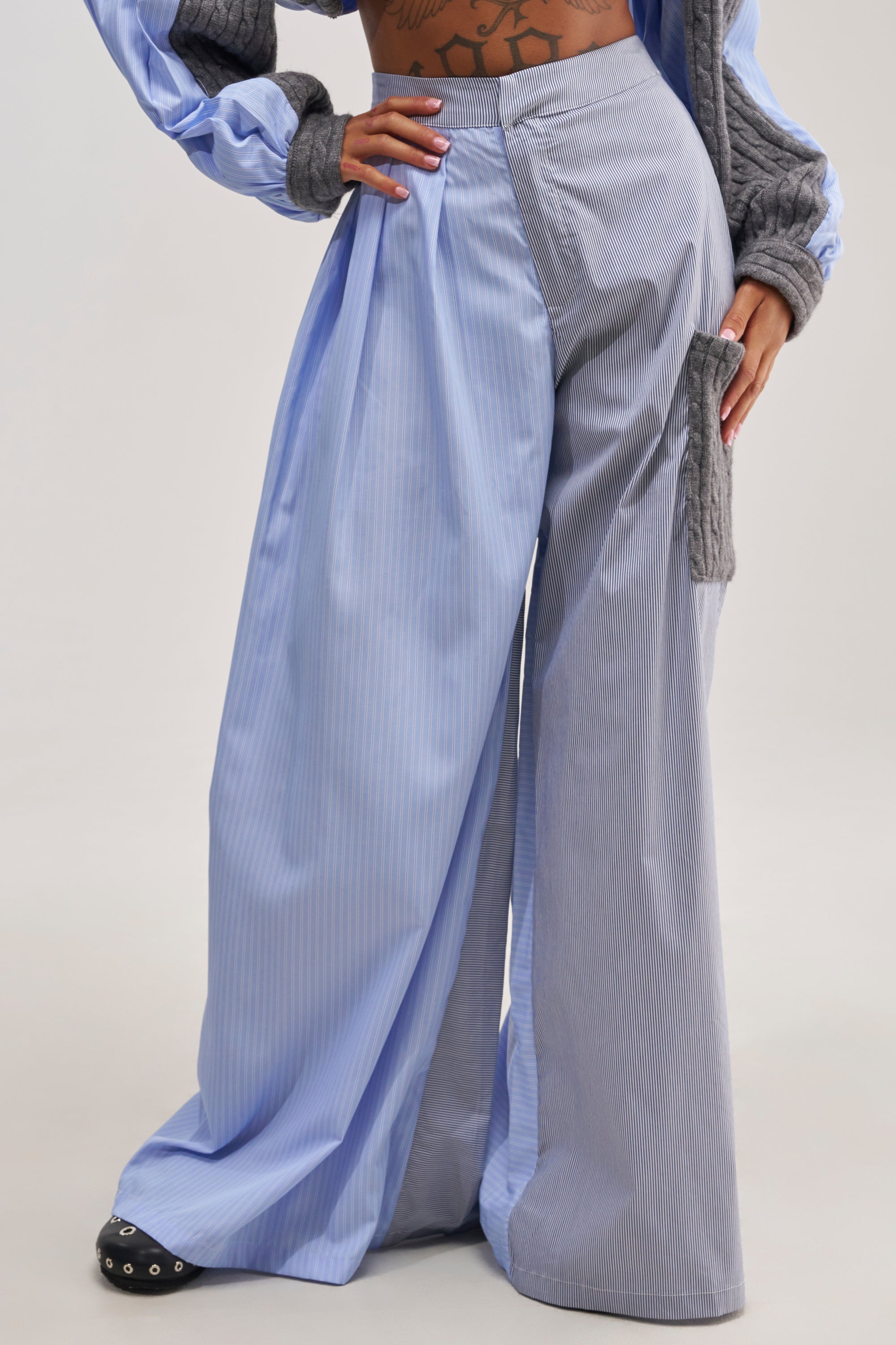 Wearing the SNUGGLES FOR EVERYONE TROUSER—colorblock wide-leg pants featuring light blue and gray pinstripes with a gray knitted pocket on one side—paired with a matching long-sleeve top. The floor-length trousers partially cover the shoes, completing a chic, fashion-forward loungewear ensemble.