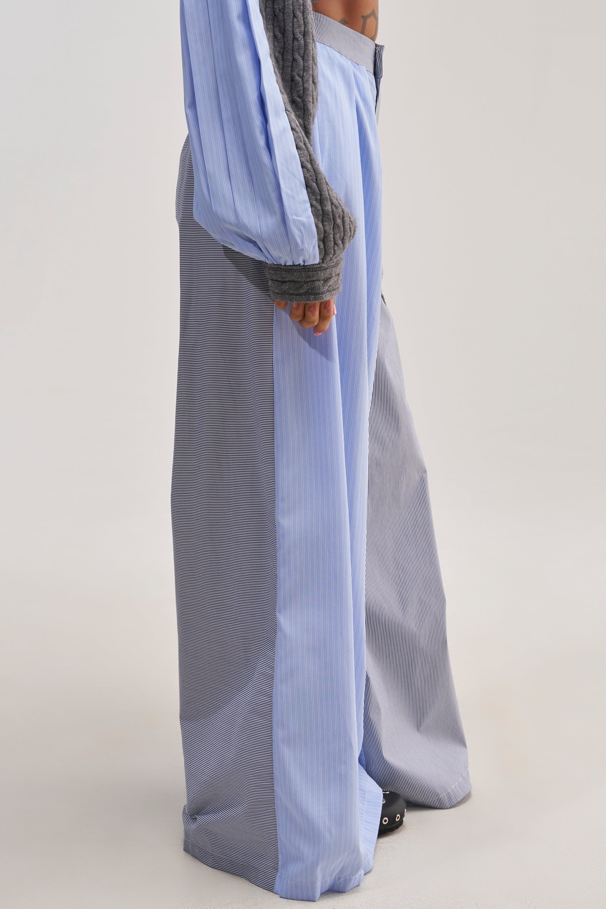 A person wearing the SNUGGLES FOR EVERYONE TROUSER—loose, wide-leg light blue cotton poplin pants with vertical stripes—paired with a gray, long-sleeve top. The image is cropped at the waist to show only the lower half of the body against a plain background.