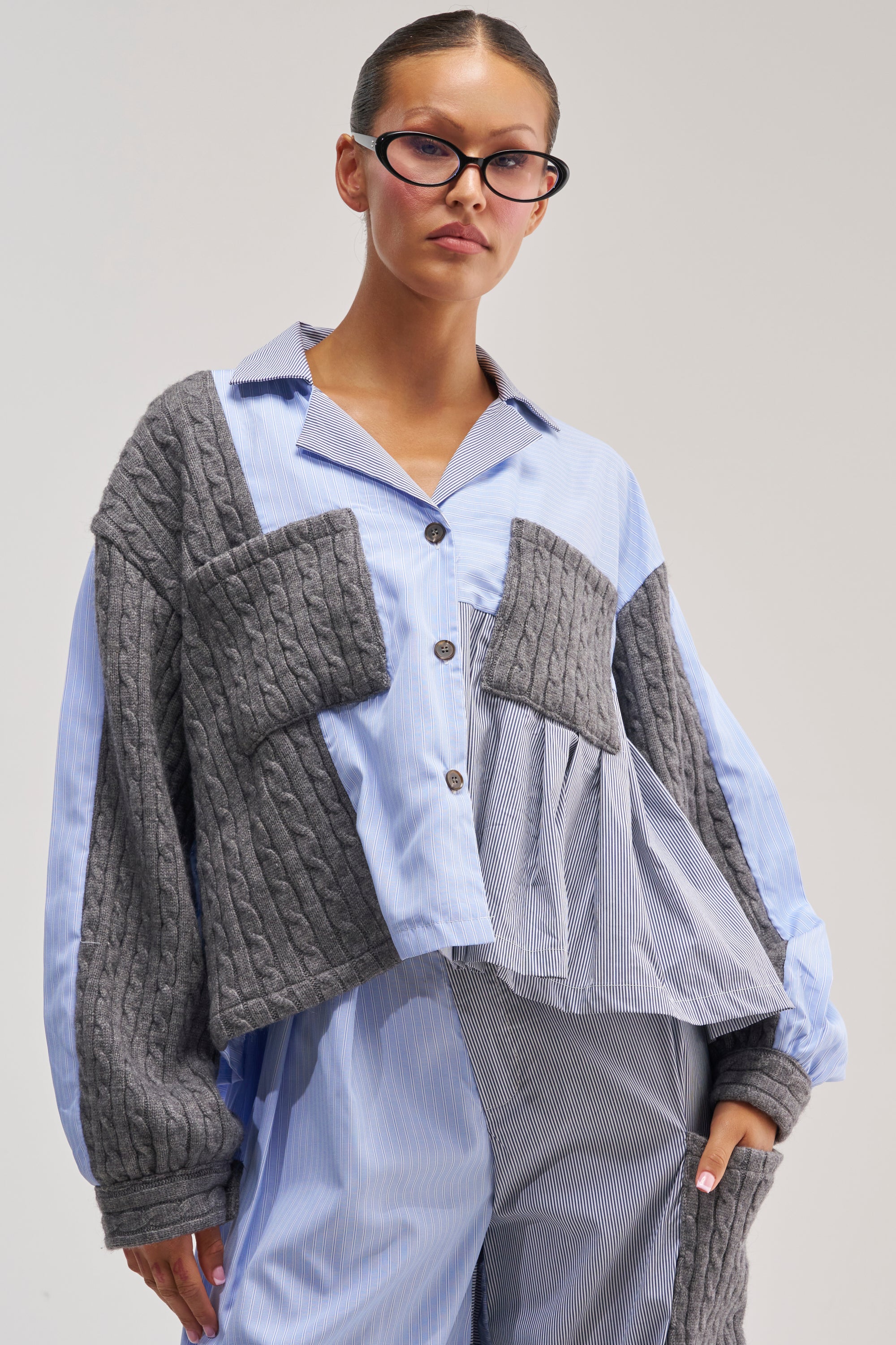 Wearing the SNUGGLES FOR EVERYONE BUTTON DOWN, a woman poses against a plain, light background. She pairs the blue striped shirt with pants and an asymmetrical gray textured patchwork sweater featuring cable-knit panels. Her look is completed with black glasses and slicked-back hair.