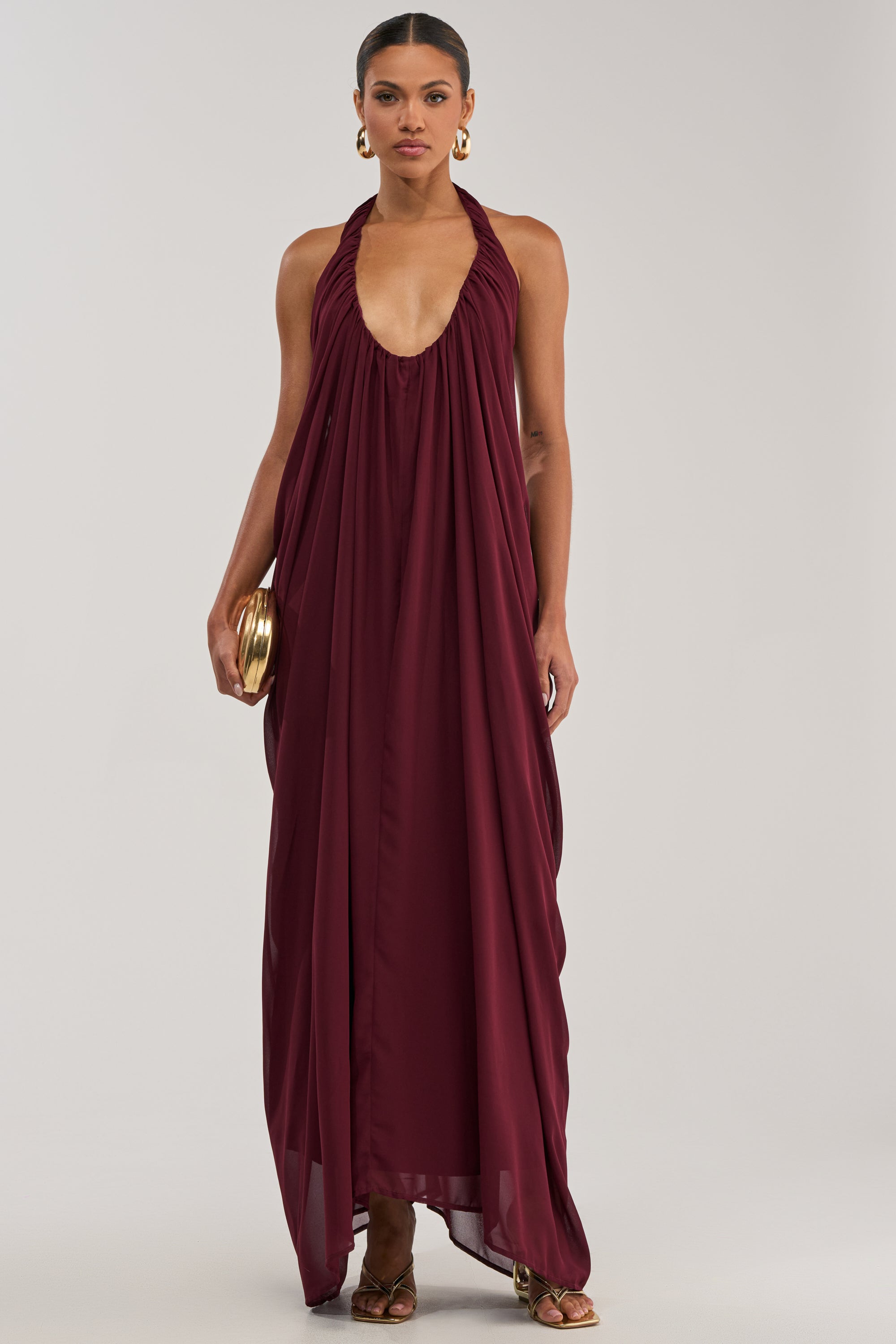 A woman stands against a plain light background wearing the RAMONA PLEATED MAXI DRESS IN BURGUNDY, styled with gold hoop earrings, gold sandals, and a small gold clutch.