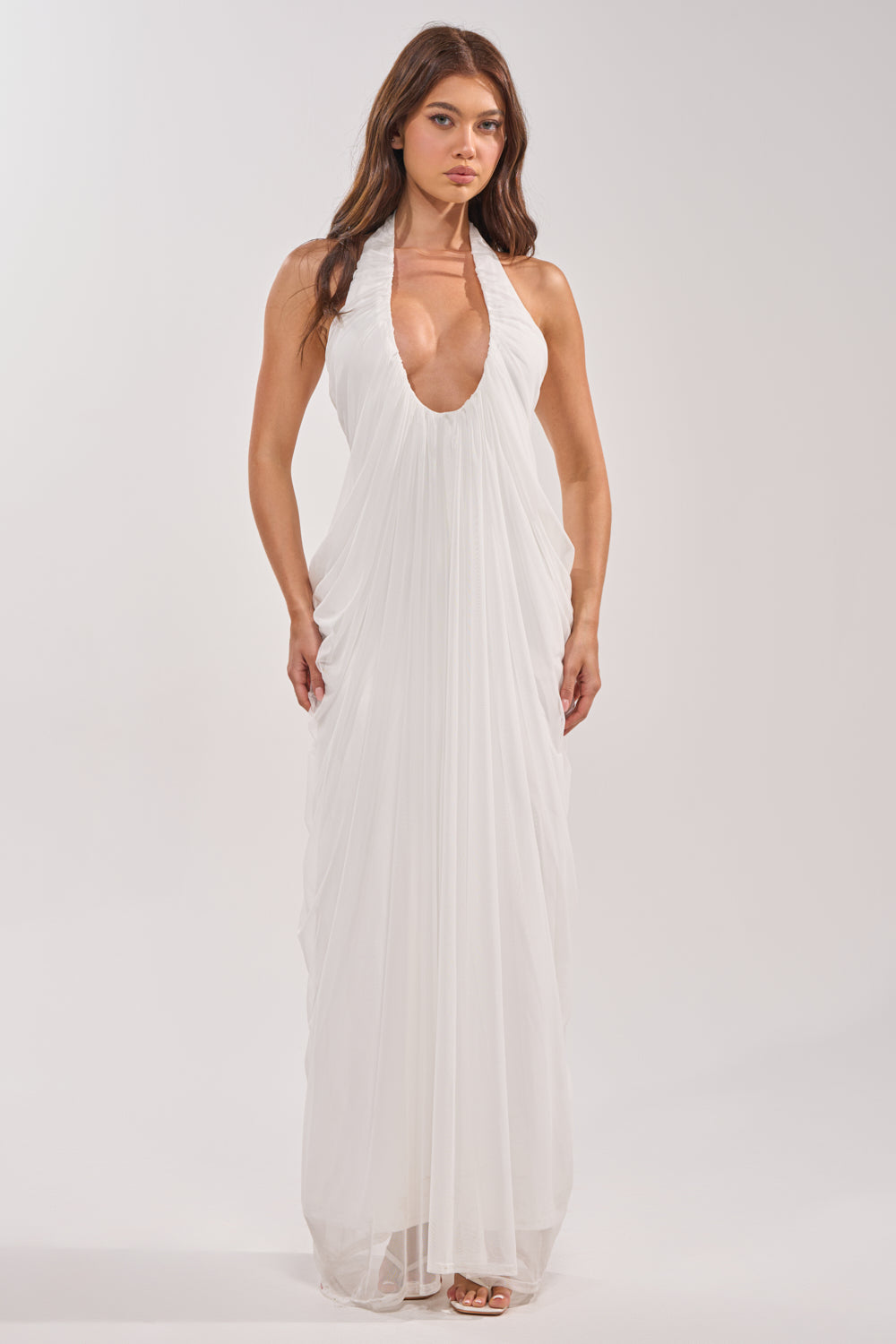 A woman wears the RAMONA PLEATED MAXI DRESS IN WHITE, featuring a halter neckline, deep plunging front, and flowing pleats. Ideal for bridal events or special occasions, she poses against a plain light background. 0