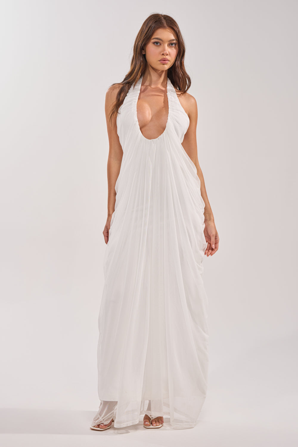 A woman with long brown hair poses against a plain background in the RAMONA PLEATED MAXI DRESS IN WHITE, a sleeveless, floor-length maxi dress featuring pleats, a plunging halter neckline, flowing draped fabric, and paired with white sandals. 1