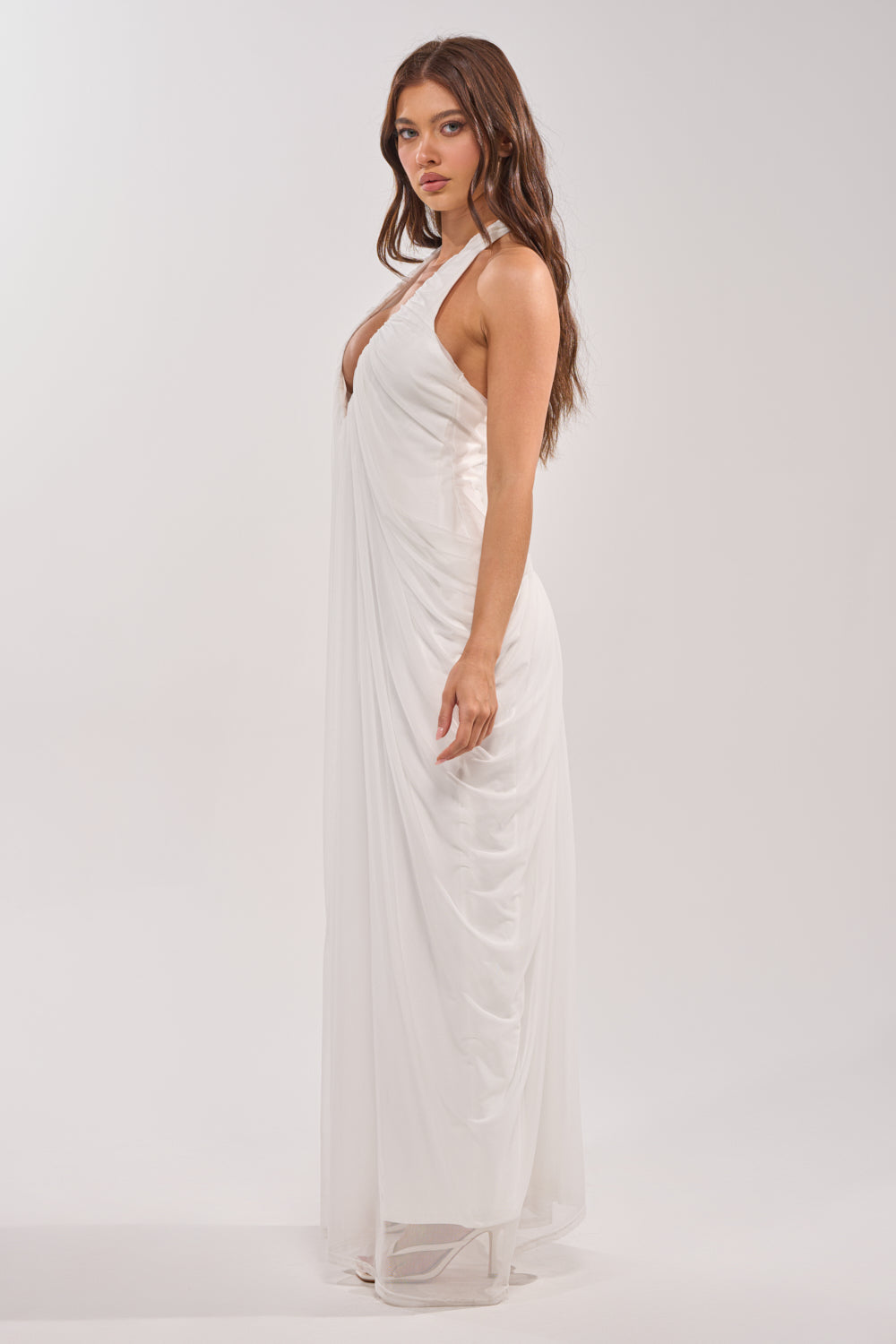 A woman with long brown hair stands in profile, wearing the RAMONA PLEATED MAXI DRESS IN WHITE, featuring a halter neckline and draped pleated fabric, against a plain light background. 2