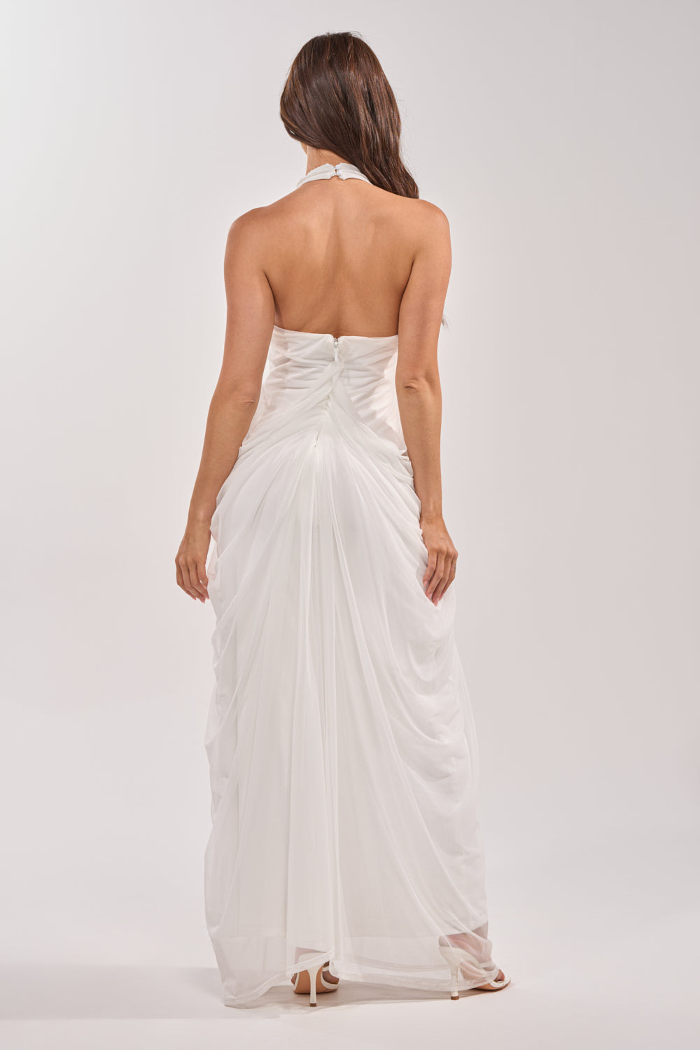 A woman with long brown hair models the RAMONA PLEATED MAXI DRESS IN WHITE, featuring a strapless, floor-length silhouette and ruched back. She faces away to showcase the elegant back details against a plain light background. 3