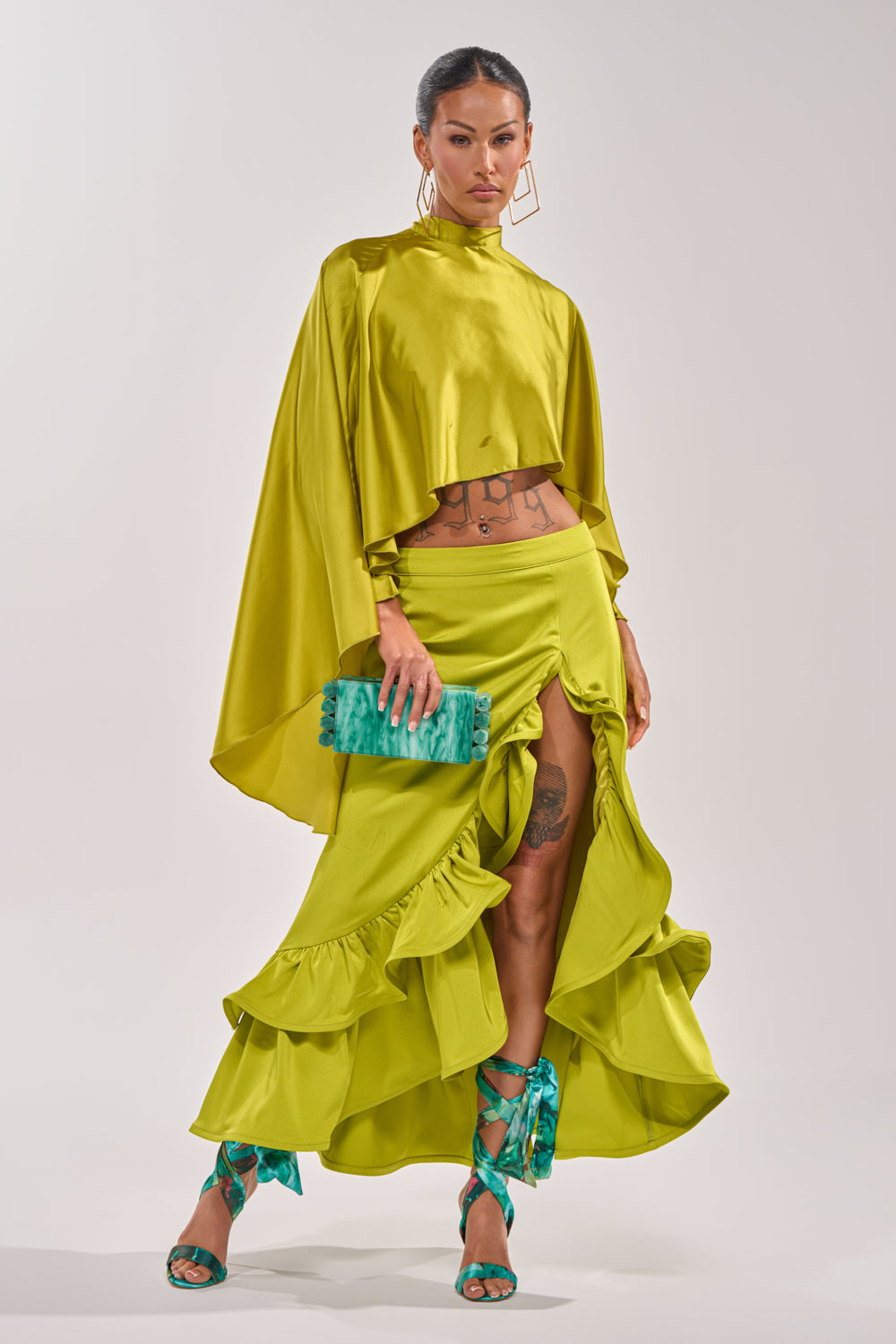 A woman models the LADY LUXE RUCHED MIDI SKIRT WITH HIGH SLIT in lime green, paired with a matching cropped top, teal clutch, teal ribbon-tied sandals, geometric earrings, and a sleek tied-back hairstyle. 0