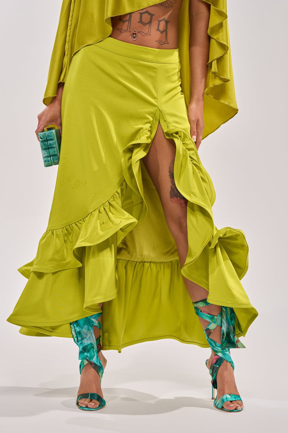 A person wears the LADY LUXE RUCHED MIDI SKIRT WITH HIGH SLIT in bright chartreuse, paired with teal strappy heels and a small teal clutch. Part of their tattooed torso is visible above the skirt. 1