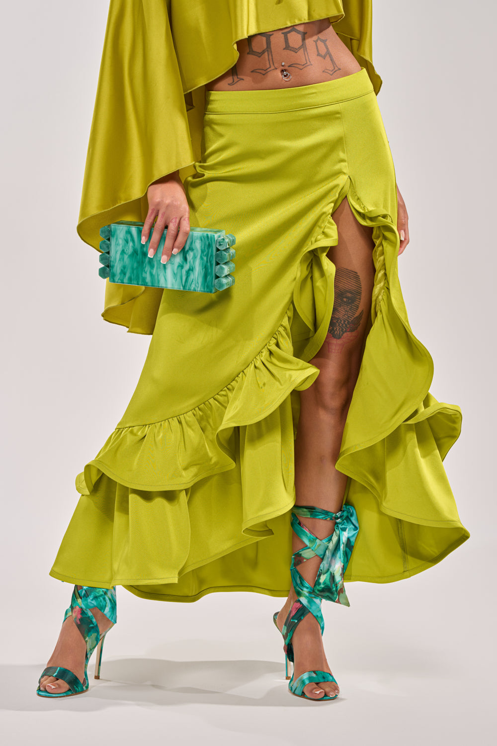 Wearing the LADY LUXE RUCHED MIDI SKIRT WITH HIGH SLIT in bright chartreuse, a person pairs it with a teal marble clutch, teal high-heeled sandals, and displays tattoo designs on their exposed skin. 2