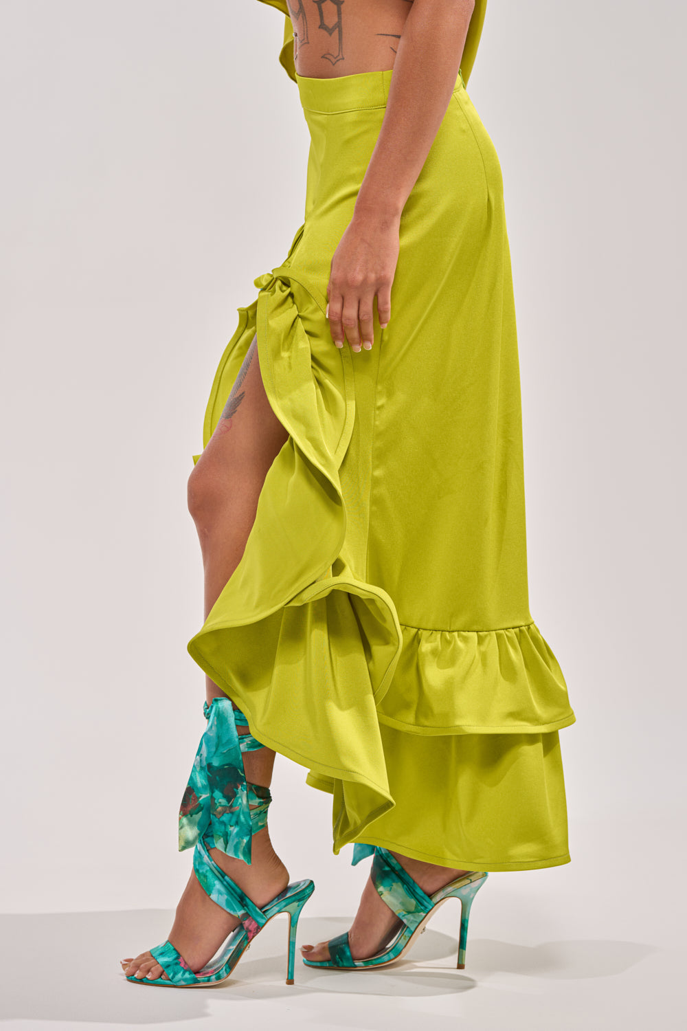 A person wears the LADY LUXE RUCHED MIDI SKIRT WITH HIGH SLIT in bright yellow-green with a high-rise waist, styled with turquoise floral high-heeled sandals featuring wrap-around ankle straps, standing against a neutral background. 3