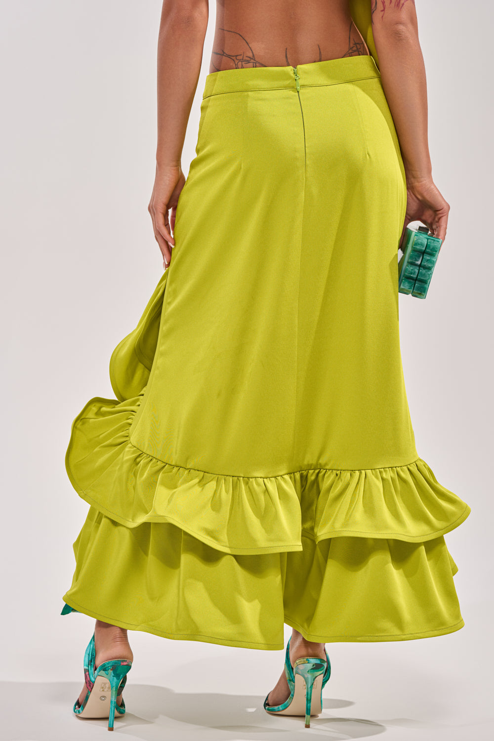 A person is seen from the back, wearing the LADY LUXE RUCHED MIDI SKIRT WITH HIGH SLIT in bright chartreuse, paired with teal high heels and a small green handbag. Their back and part of a tattoo are visible. 5