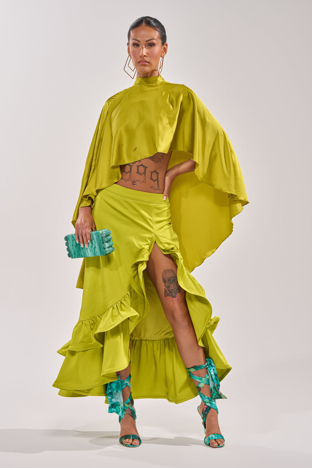 A woman showcases the LADY LUXE RUCHED MIDI SKIRT WITH HIGH SLIT in vibrant yellow-green, paired with a matching cape-style top, teal lace-up heels, large gold earrings, a textured teal clutch, and visible midriff tattoos. 6