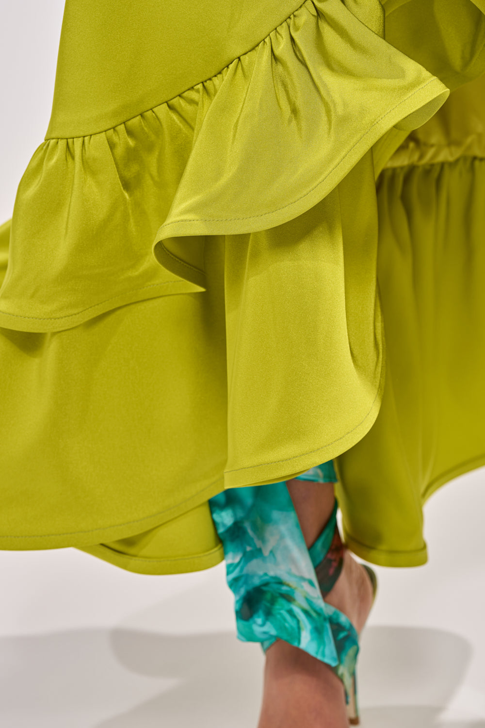 Close-up of the LADY LUXE RUCHED MIDI SKIRT WITH HIGH SLIT in chartreuse, featuring flowy ruffles and a high slit, with glimpses of turquoise patterned fabric and a high-heeled sandal underneath. 8