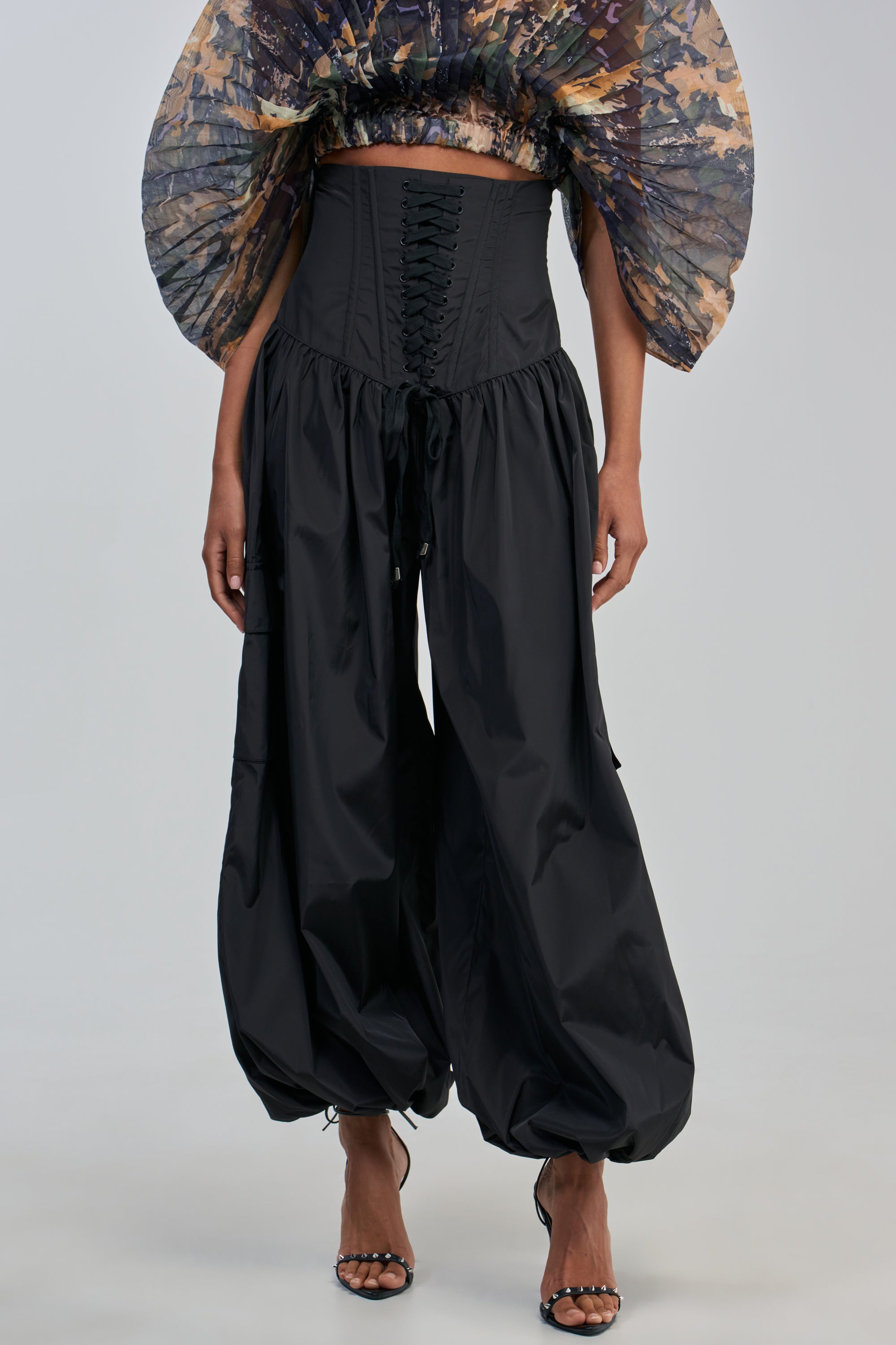 A person wearing the LOCKED UP FOR GOOD BALLOON PANT IN BLACK, featuring black lace-up corset detail at the waist and wide, gathered legs reminiscent of balloon pants. Only the lower half and part of the torso are visible. The look is completed with black heeled sandals and a sheer, patterned, ruffled top, creating a bold streetwear statement.