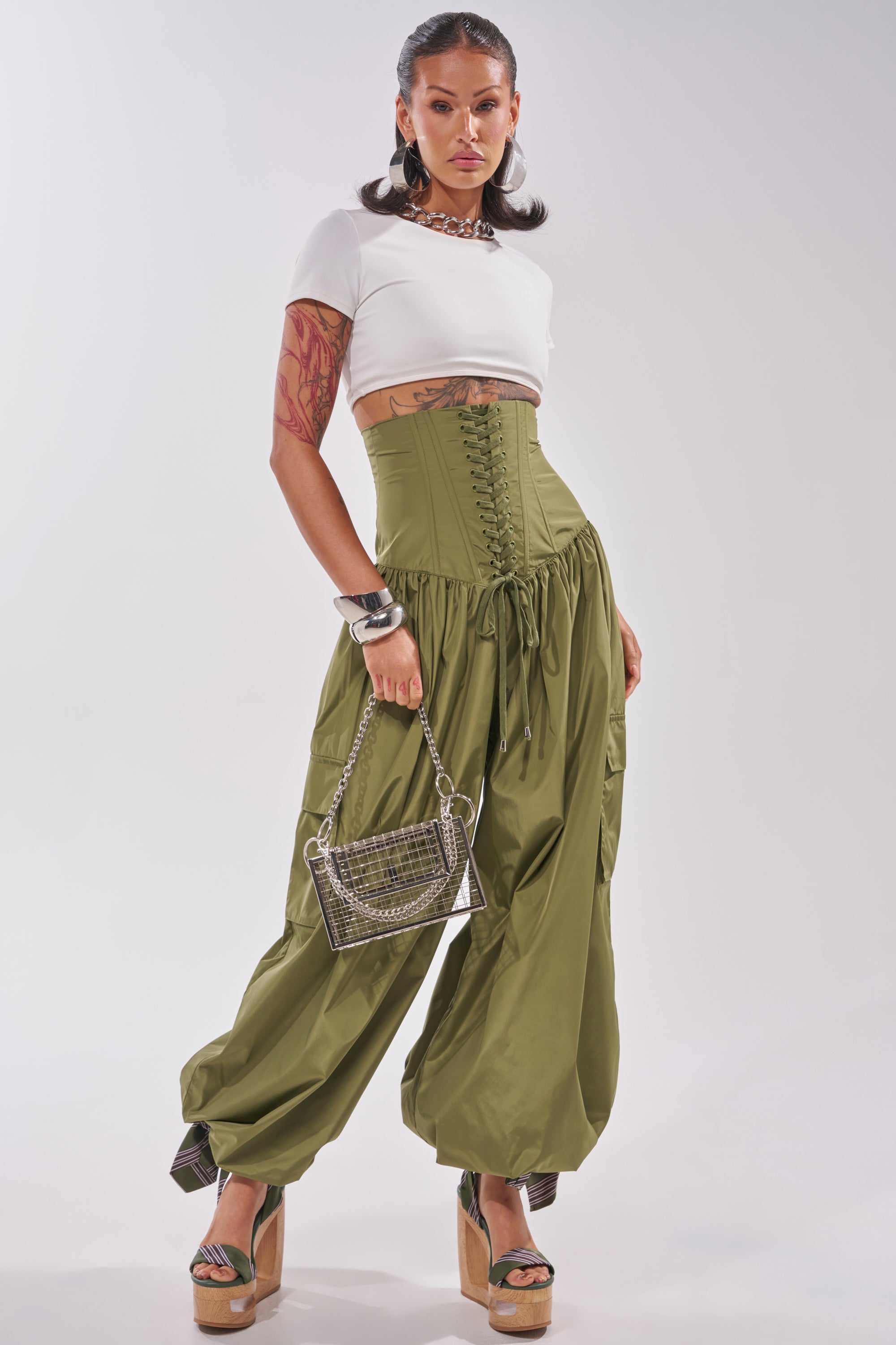 A woman poses in a white crop top and the LOCKED UP FOR GOOD LACE UP BALLOON PANT, styled with platform heels, a metallic purse, bold jewelry, and showing off her arm tattoos. 0
