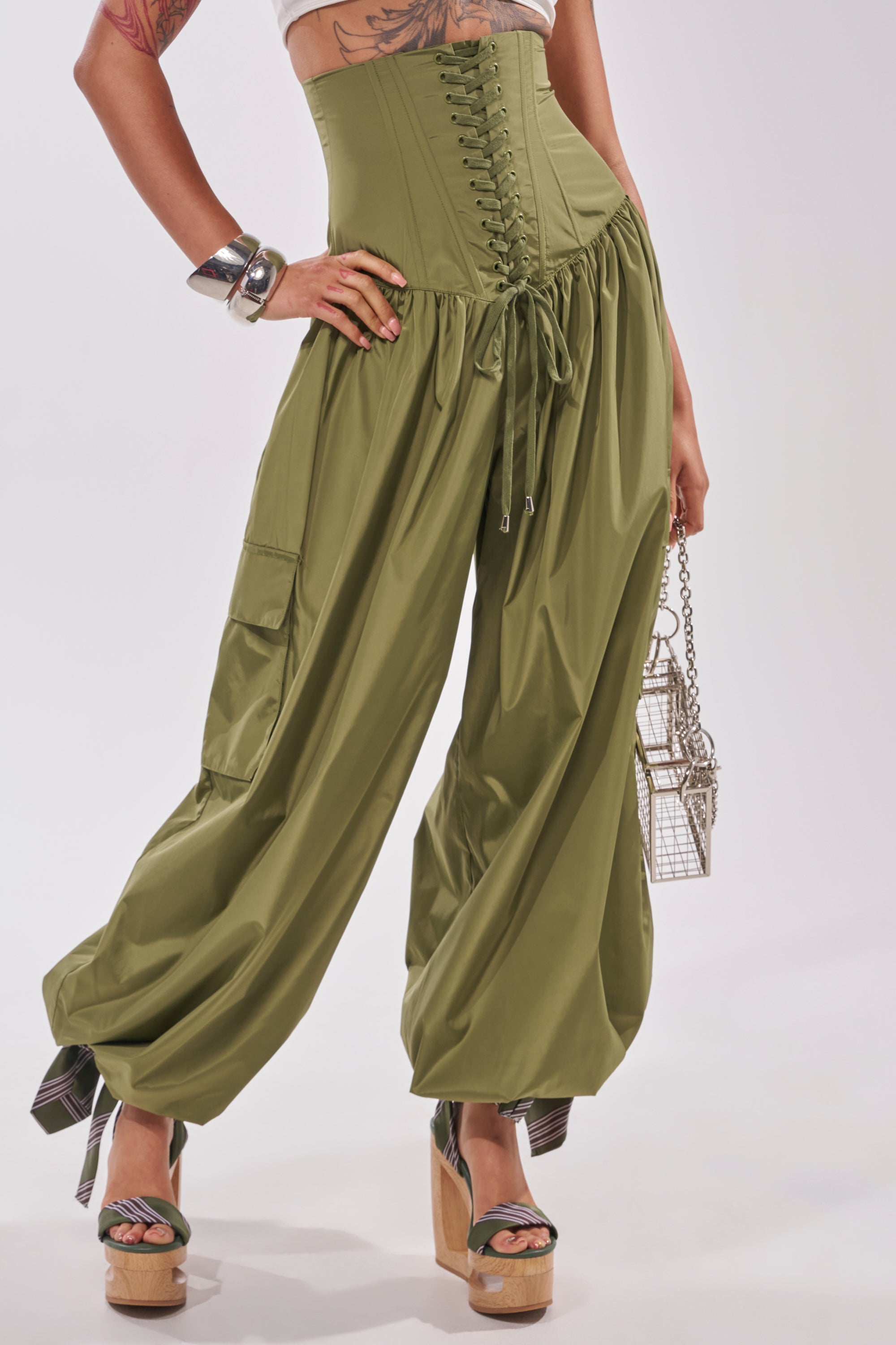 A person wears the LOCKED UP FOR GOOD LACE UP BALLOON PANT in olive green with lace-up balloon legs, styled with platform sandals, silver bangles, and a small metallic handbag—capturing a trendy festival vibe. The top of the head is not shown. 1