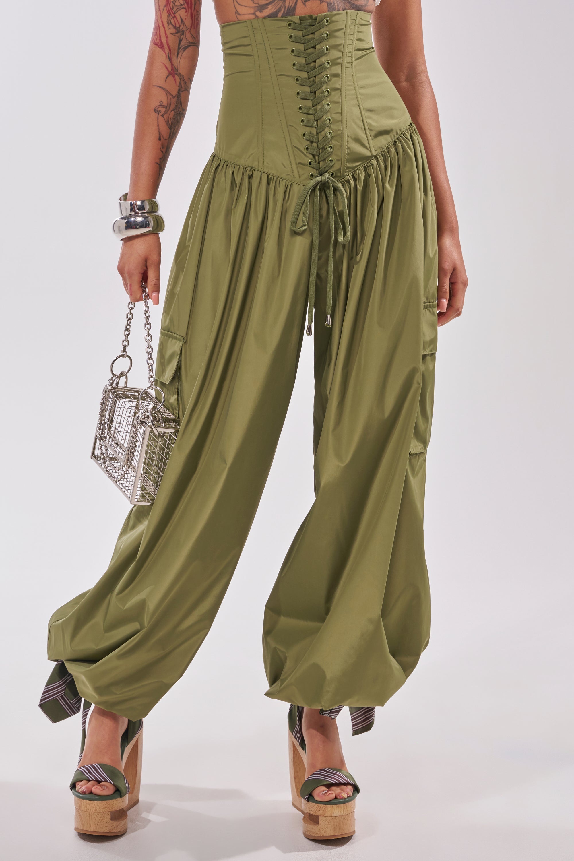 Wearing the LOCKED UP FOR GOOD LACE UP BALLOON PANT in olive green, styled with a corset-style front, wooden platform sandals, silver mesh purse, chunky bracelets, and visible arm tattoos. 2