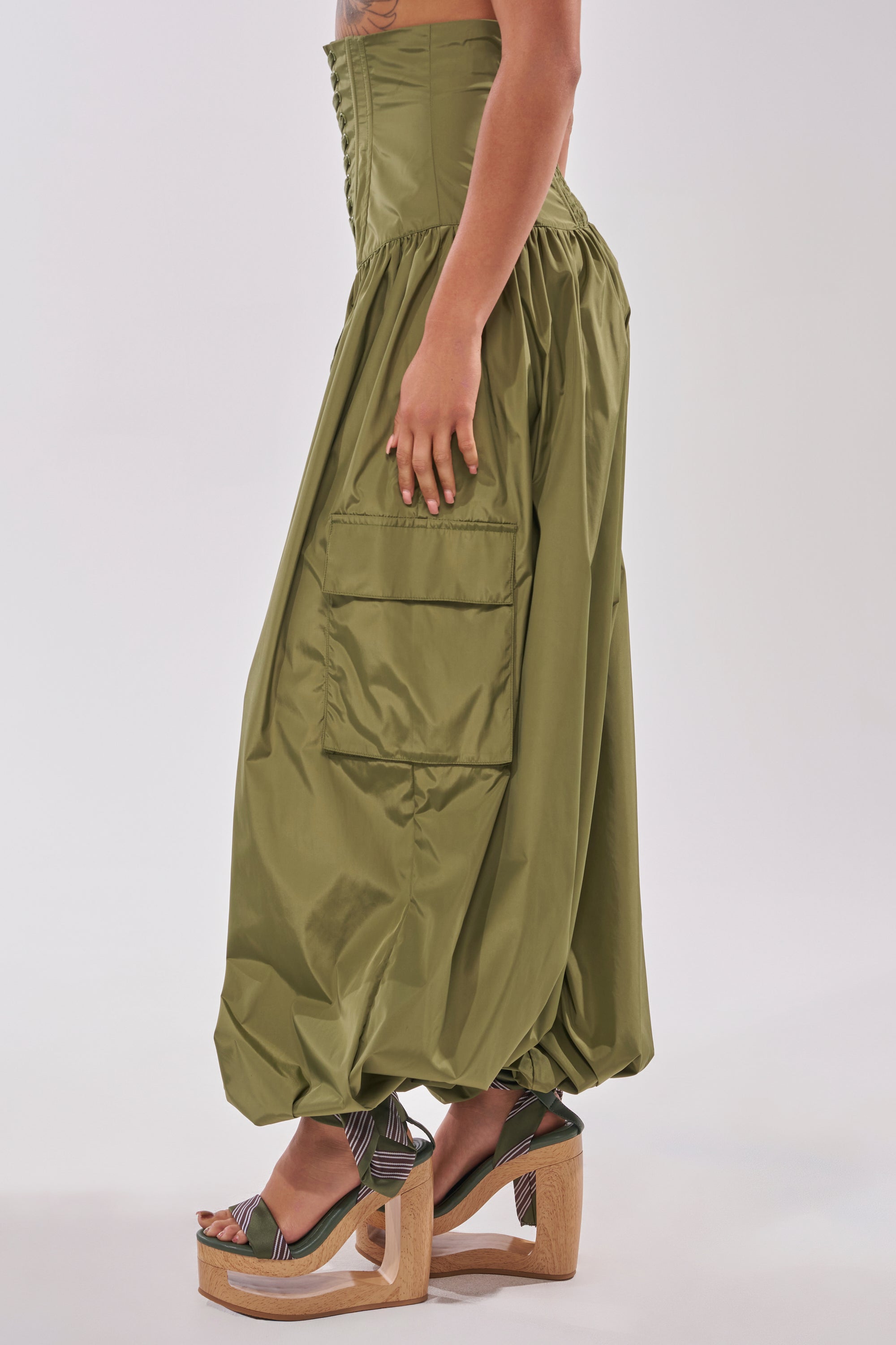 A person wears the LOCKED UP FOR GOOD LACE UP BALLOON PANT in olive green, featuring high waist, large side pockets, and elastic cuffs, paired with platform wedge sandals against a light background. 3