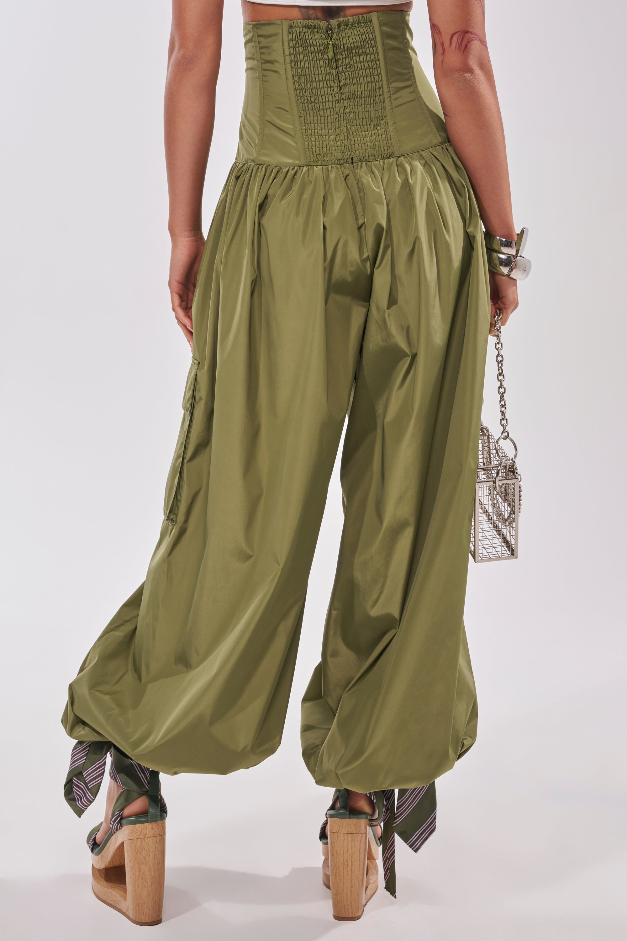 Someone wearing the LOCKED UP FOR GOOD LACE UP BALLOON PANT with a corset-style top and wooden platform sandals, holding a small silver cage purse, is photographed from behind. 4