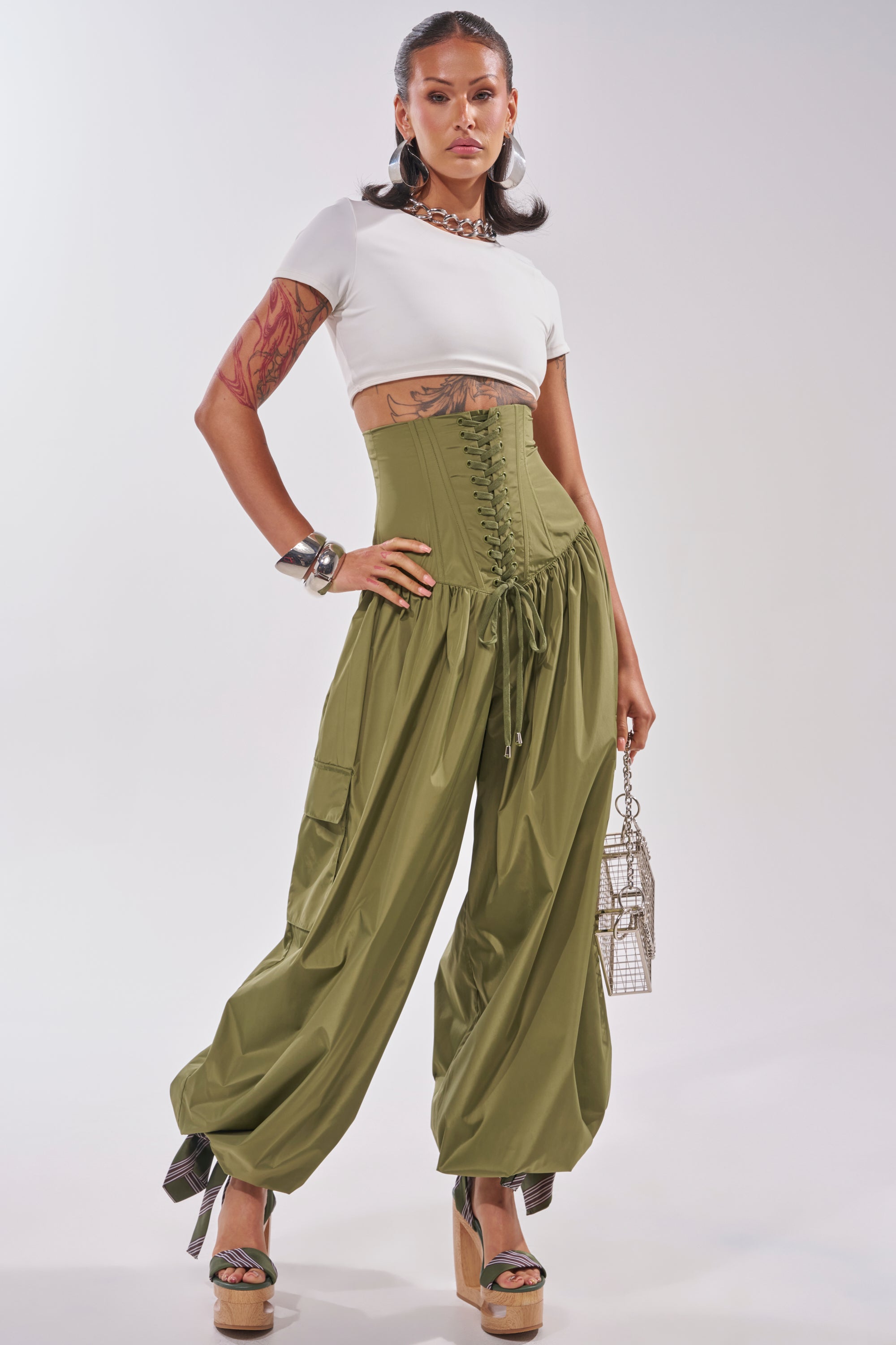 A person poses confidently in fitted white crop top, platform heels, jewelry, metallic handbag, and LOCKED UP FOR GOOD LACE UP BALLOON PANT featuring an olive corset waist and balloon legs. 5
