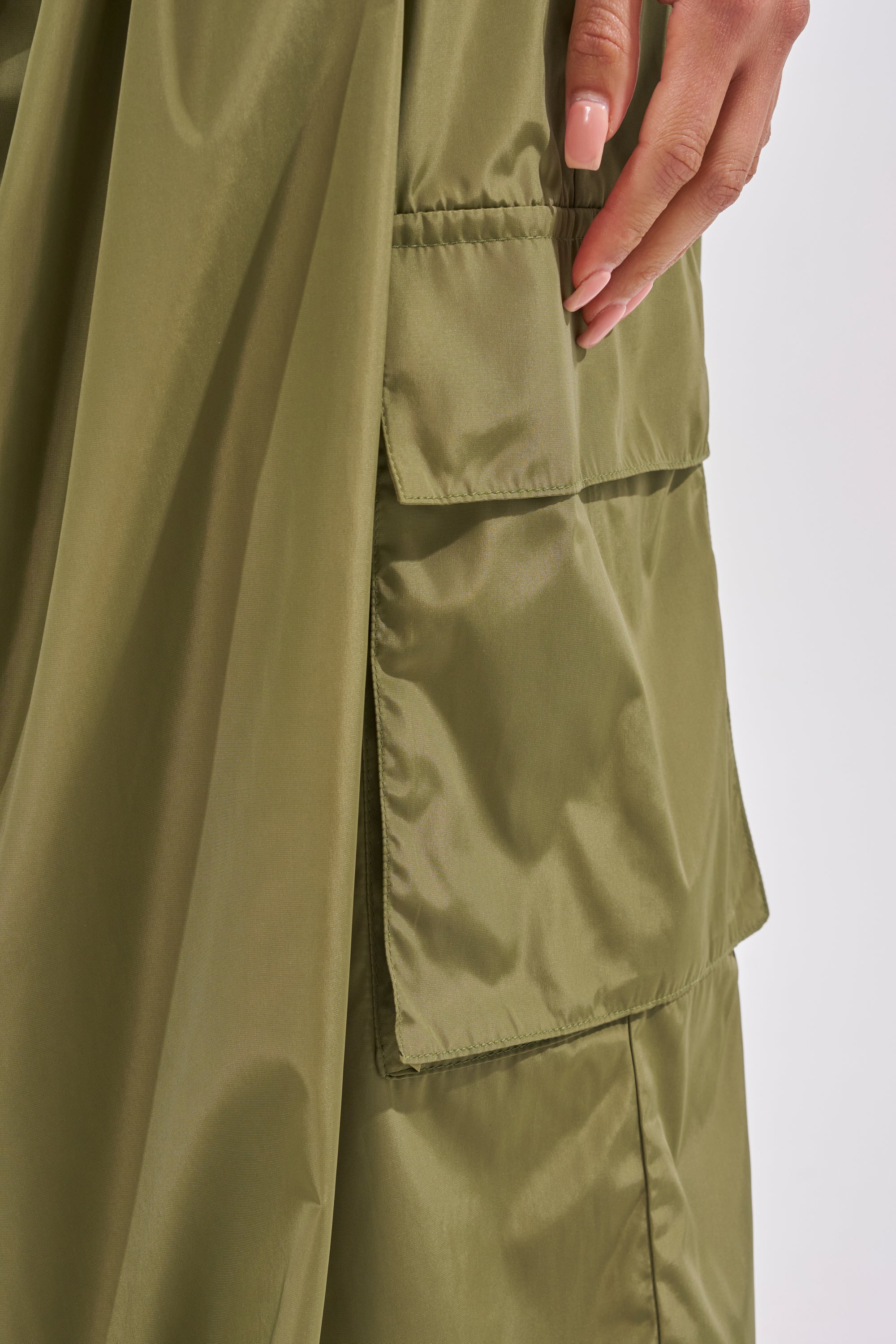 A close-up of a manicured hand resting next to a large olive green pocket on the shiny LOCKED UP FOR GOOD LACE UP BALLOON PANT. 7