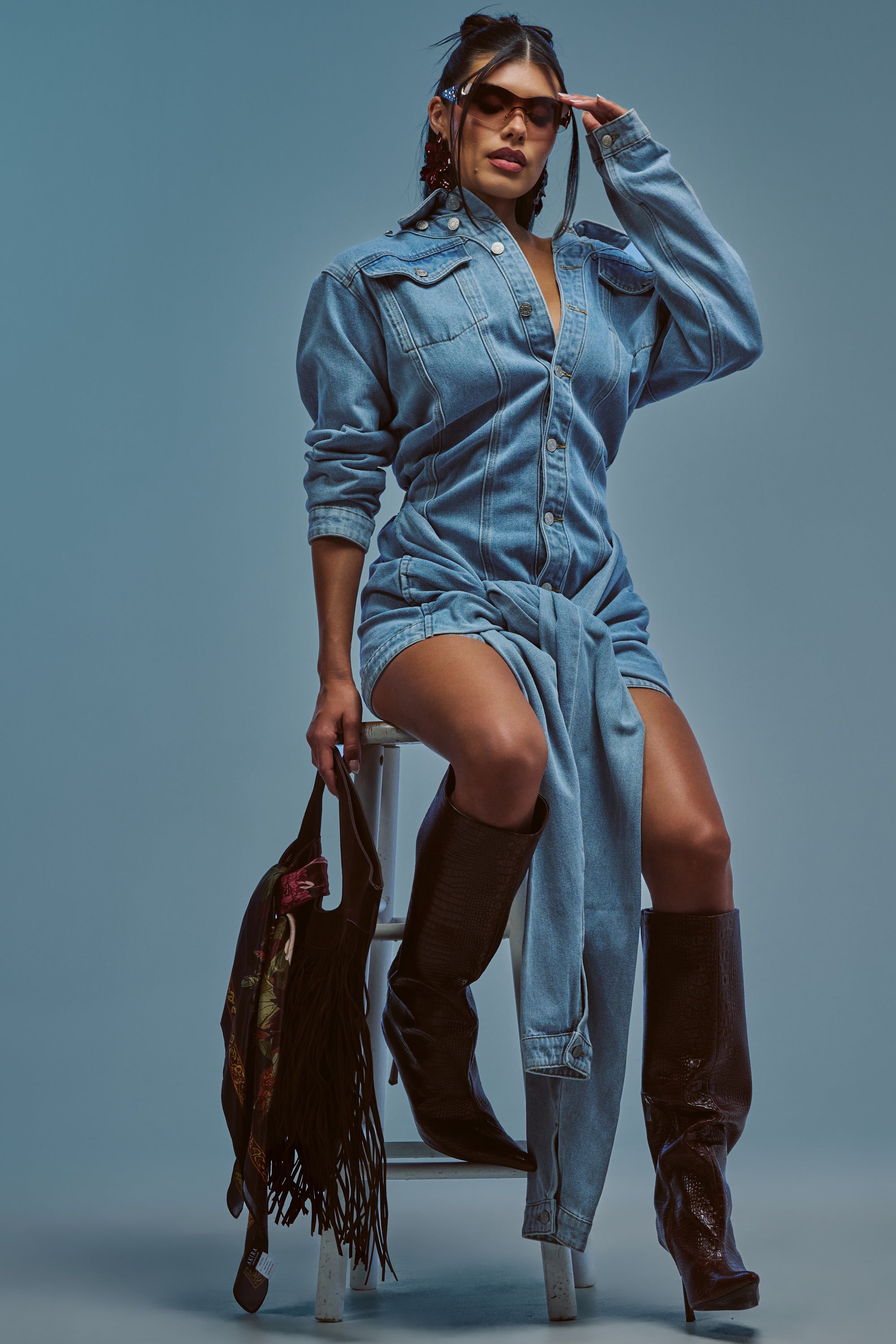 Wearing the MAGNETIC ENERGY DENIM MINI DRESS, a person sits on a stool against a blue background. They accessorize with sunglasses, black boots, a fringed bag, and have another denim shirt tied around their waist.
