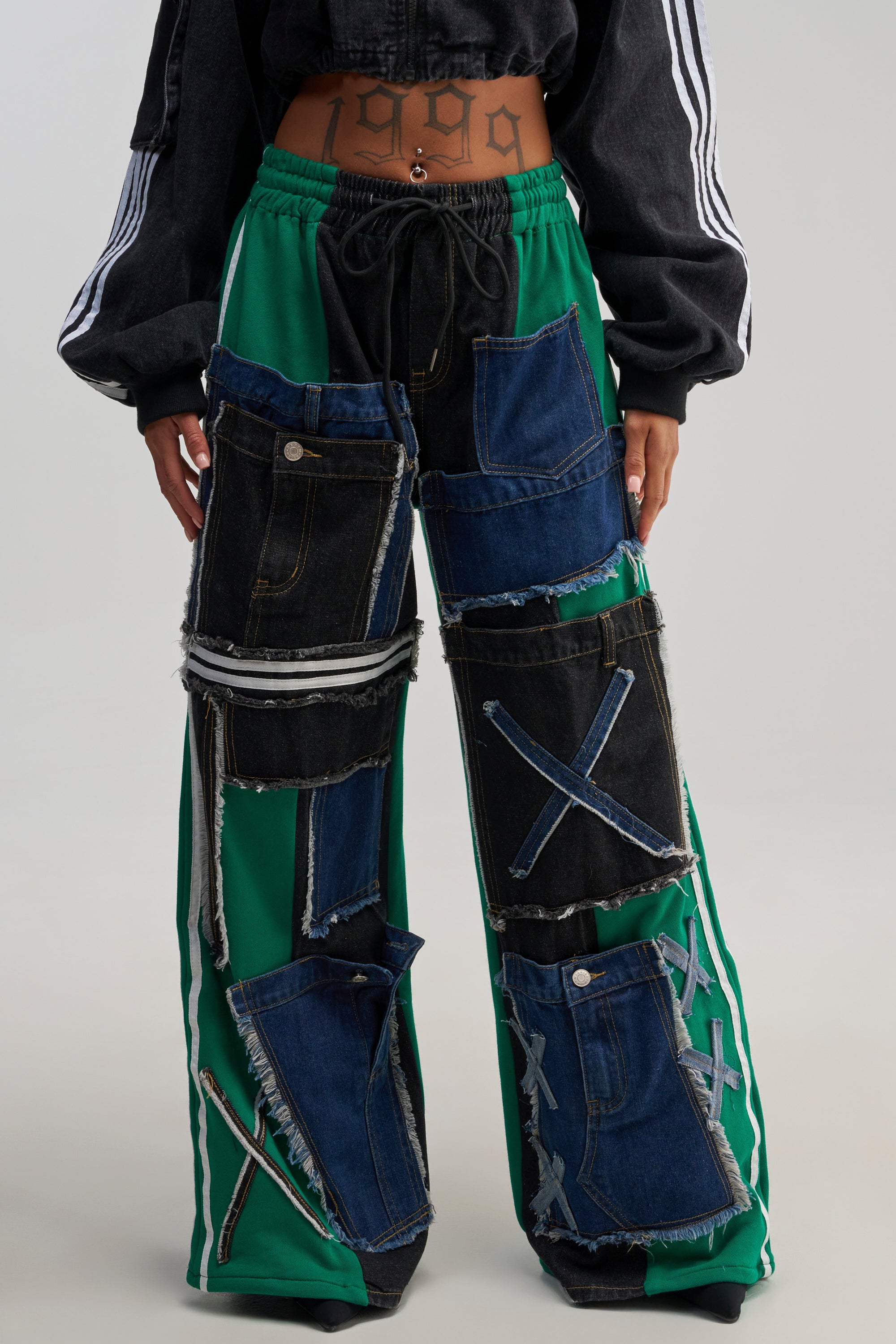 Wearing the GAME DAY VIBES JOGGER—green wide-leg joggers featuring patchwork denim details and distinctive pockets—a person pairs them with a cropped black jacket, showcasing a visible “1999” tattoo on their midriff.