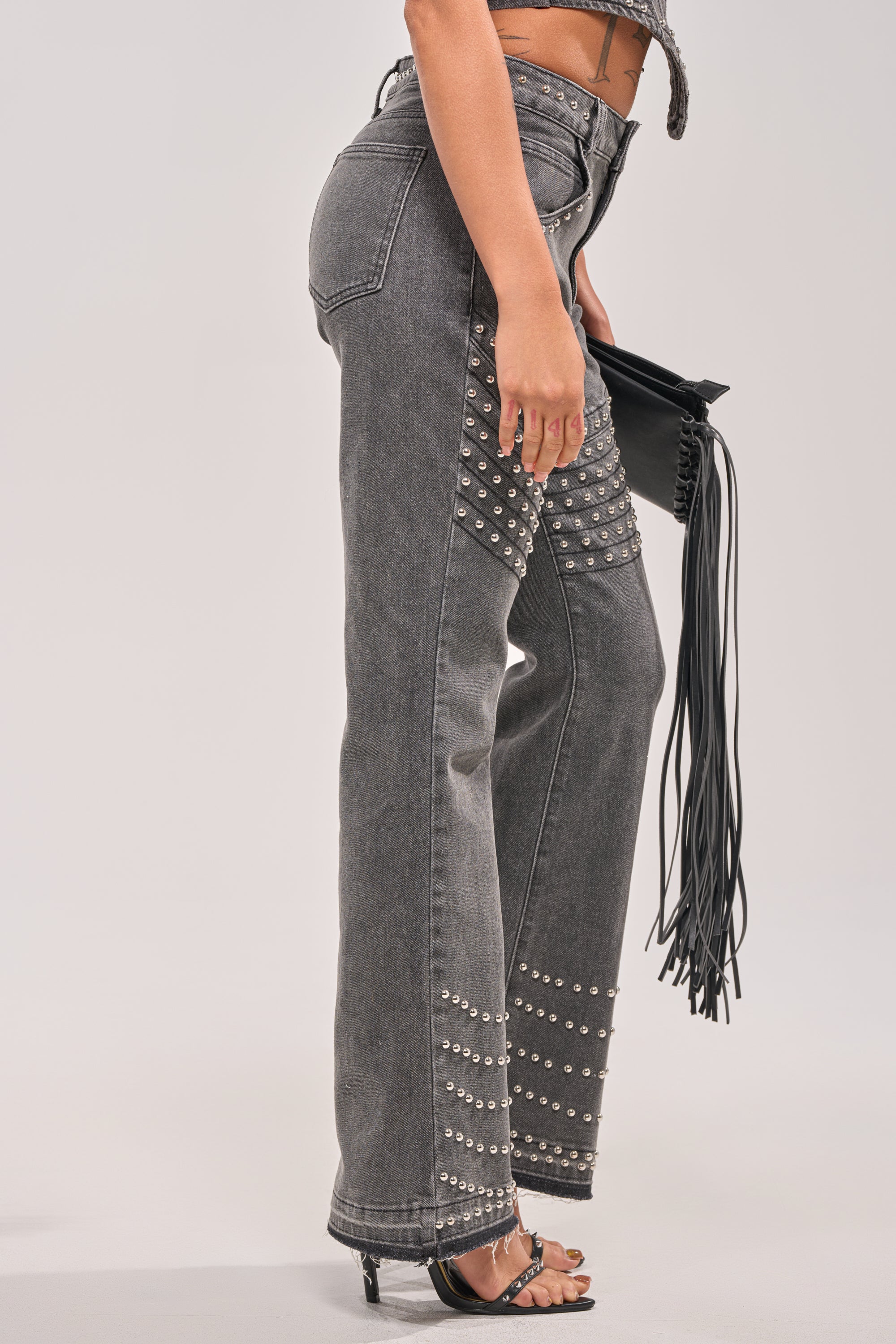 A person wearing FEELING THE VIBES FLARE DENIM—gray high-waisted, straight-leg jeans with silver dome studs—paired with black heeled sandals and a black fringe clutch bag for a rocker-chic look. 2