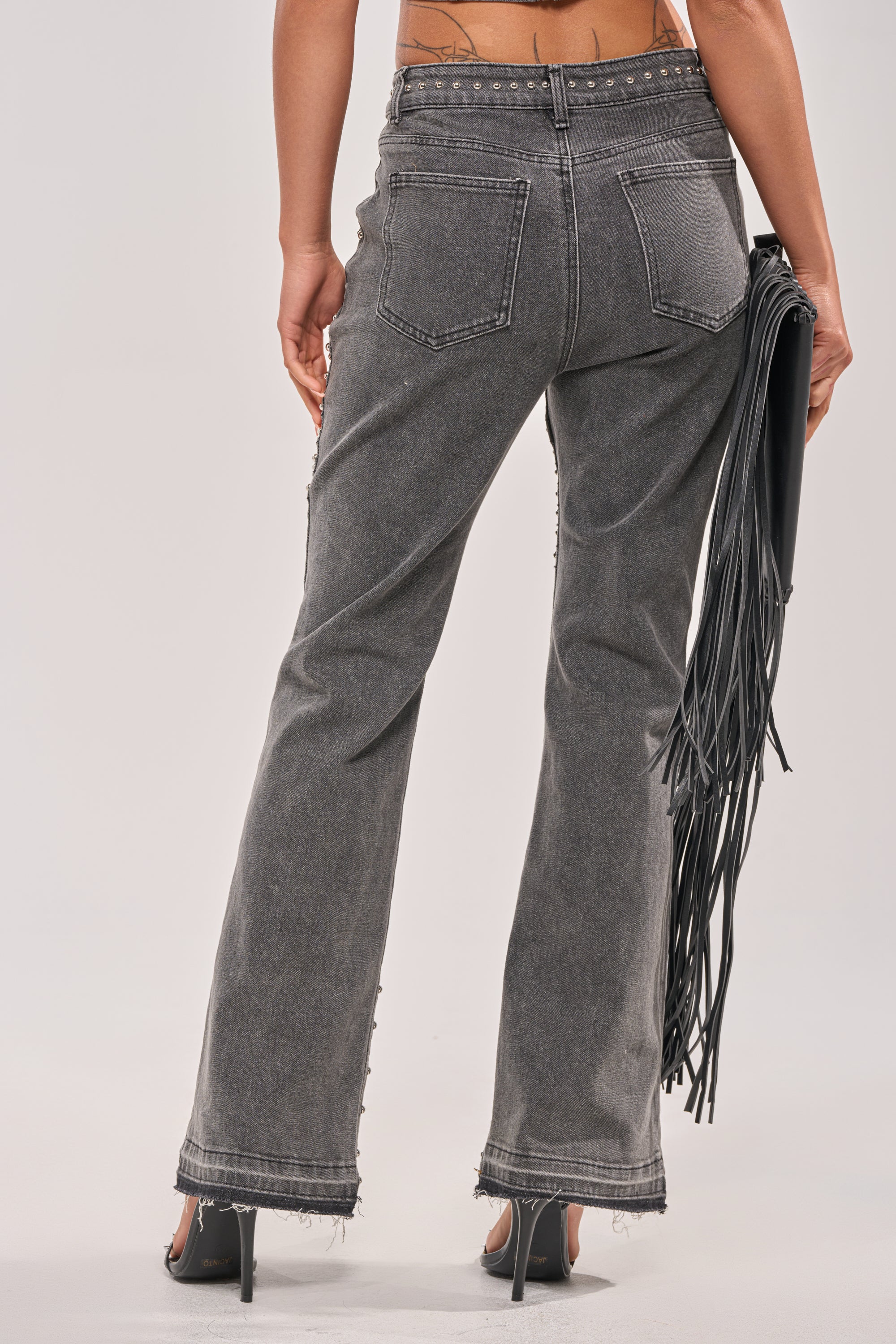 A person stands with their back to the camera, wearing FEELING THE VIBES FLARE DENIM jeans with raw hems, black heels, and holding a black fringed bag in their right hand against a plain light background. 3