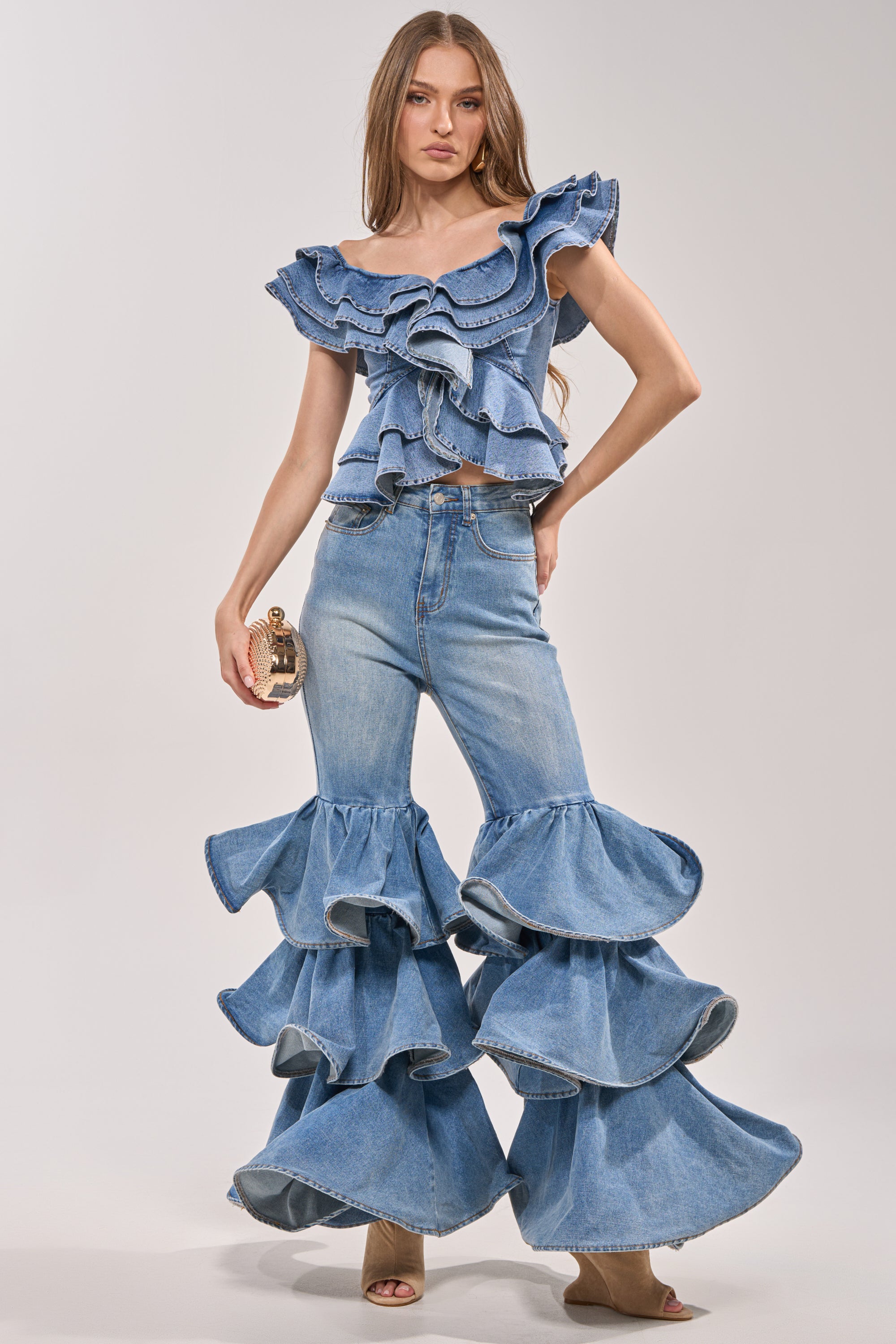 A woman models the RUFFLE CITY DENIM: a ruffled denim crop top with matching western-inspired jeans featuring oversized, layered ruffles below the knees. She holds a small clutch against a plain, light backdrop. 0