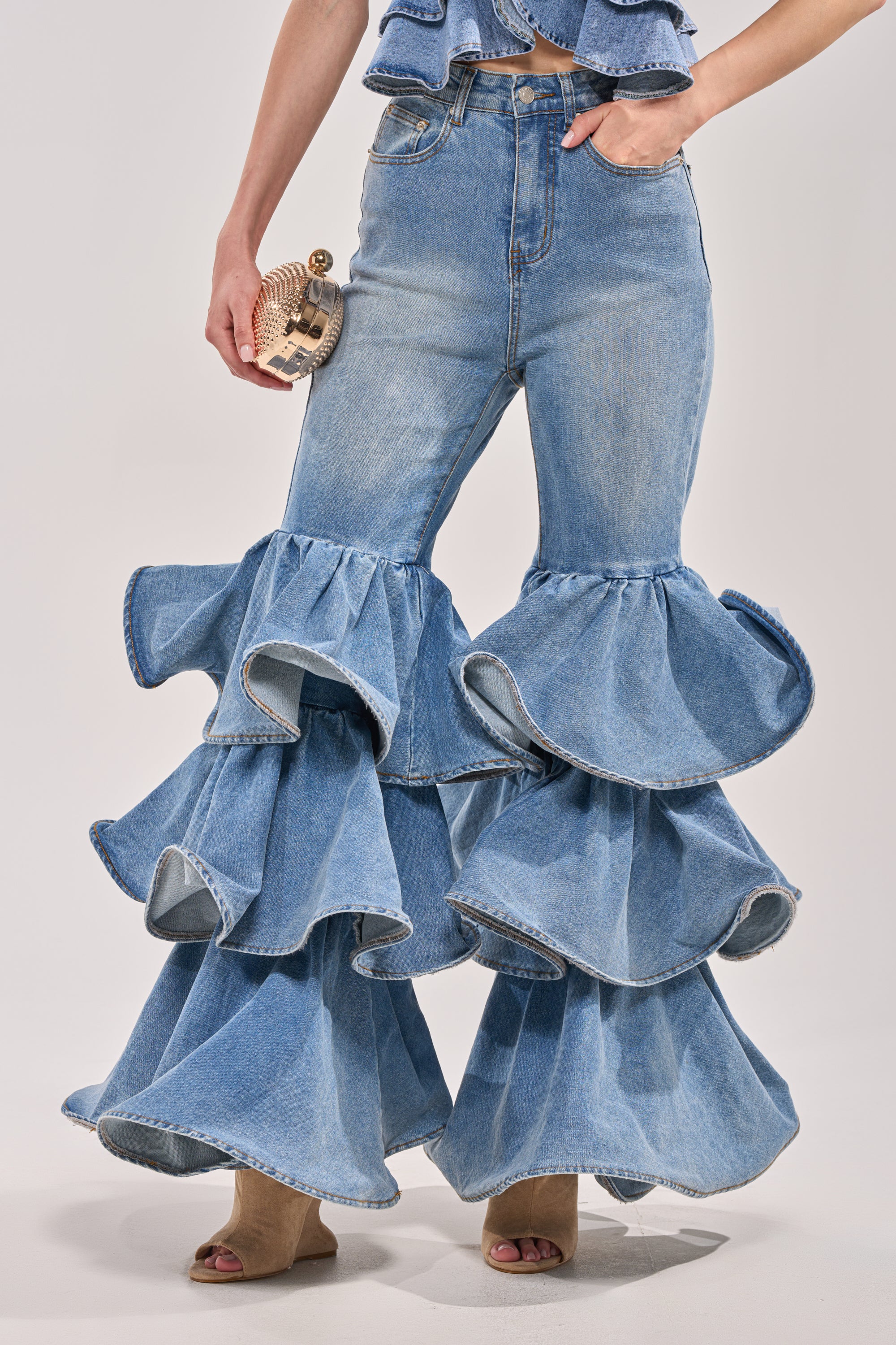 Wearing RUFFLE CITY DENIM high-rise stretch jeans featuring dramatic layered ruffles at the knees and flared legs, paired with open-toe heels and a small round clutch. 1