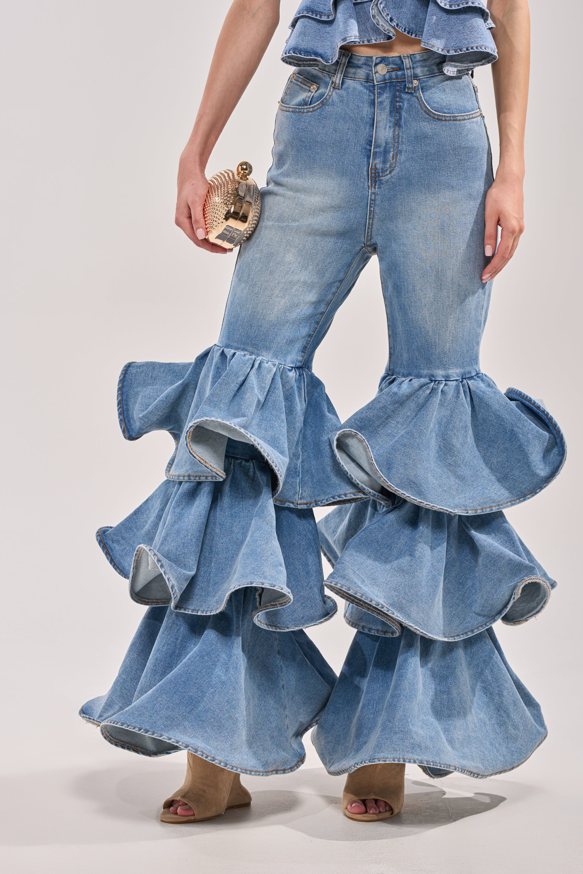 Wearing RUFFLE CITY DENIM—light blue ruffle flare jeans with large layered ruffles from the knees down—styled with a small clutch and open-toe heels, these western-inspired jeans pop against a plain light background. 2