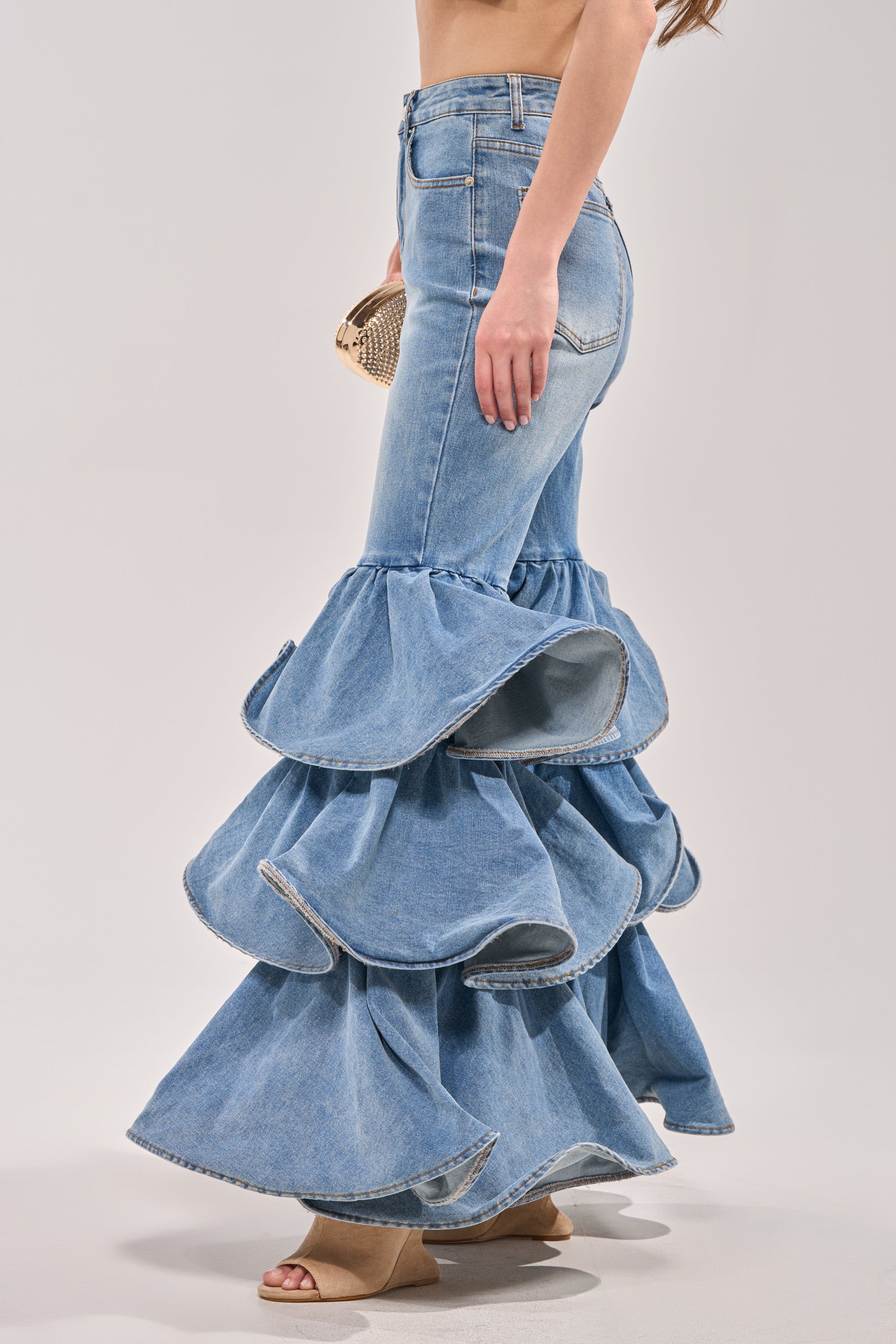 Wearing the RUFFLE CITY DENIM strapless dress with dramatic tiered ruffles, a woman pairs it with a small clutch and beige open-toe heels, posing against a plain light background. 3