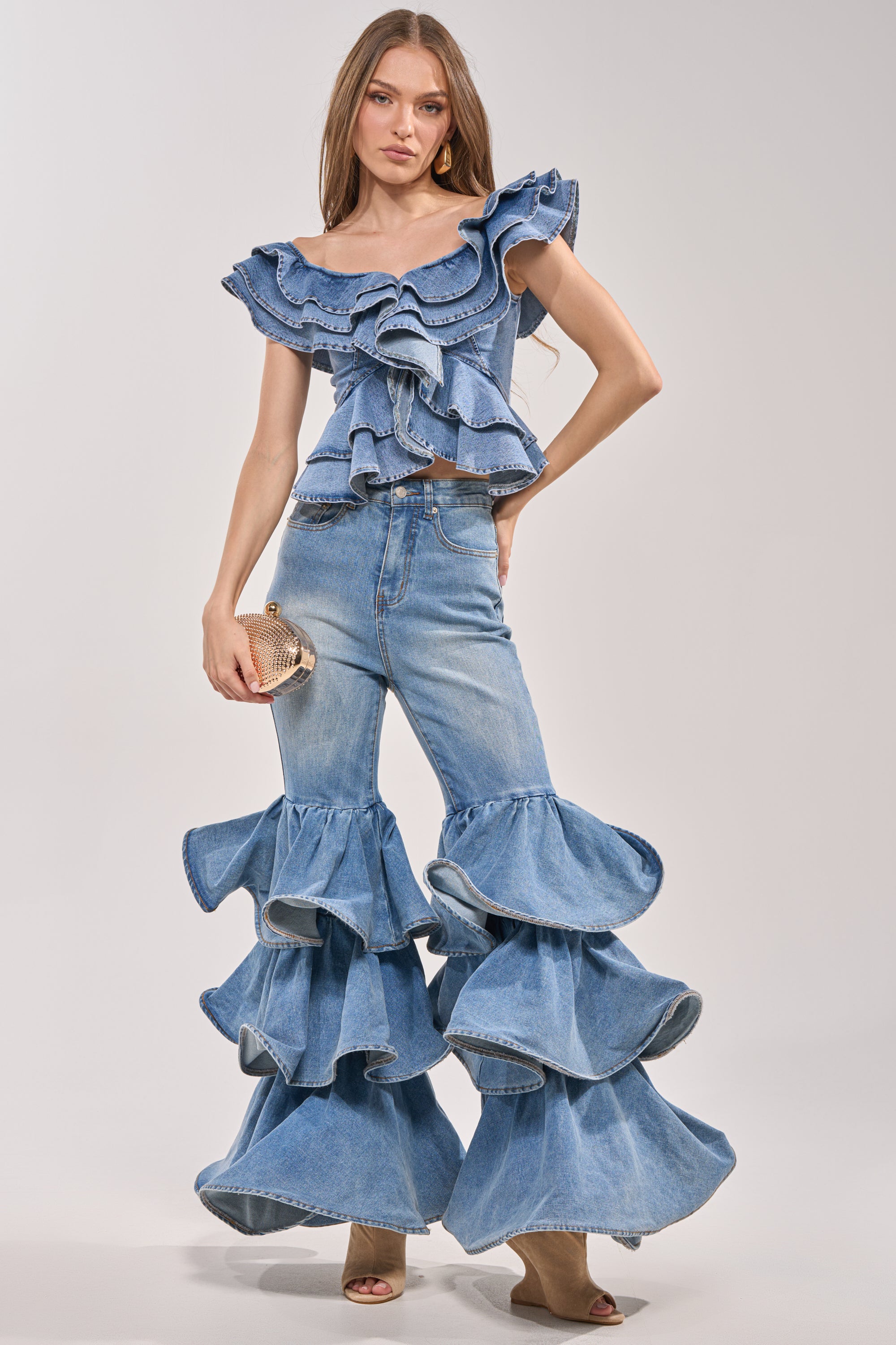A woman poses in RUFFLE CITY DENIM high-rise stretch flare jeans with ruffle legs and a matching ruffled top, holding a beige clutch and wearing sandals against a plain light background. 5