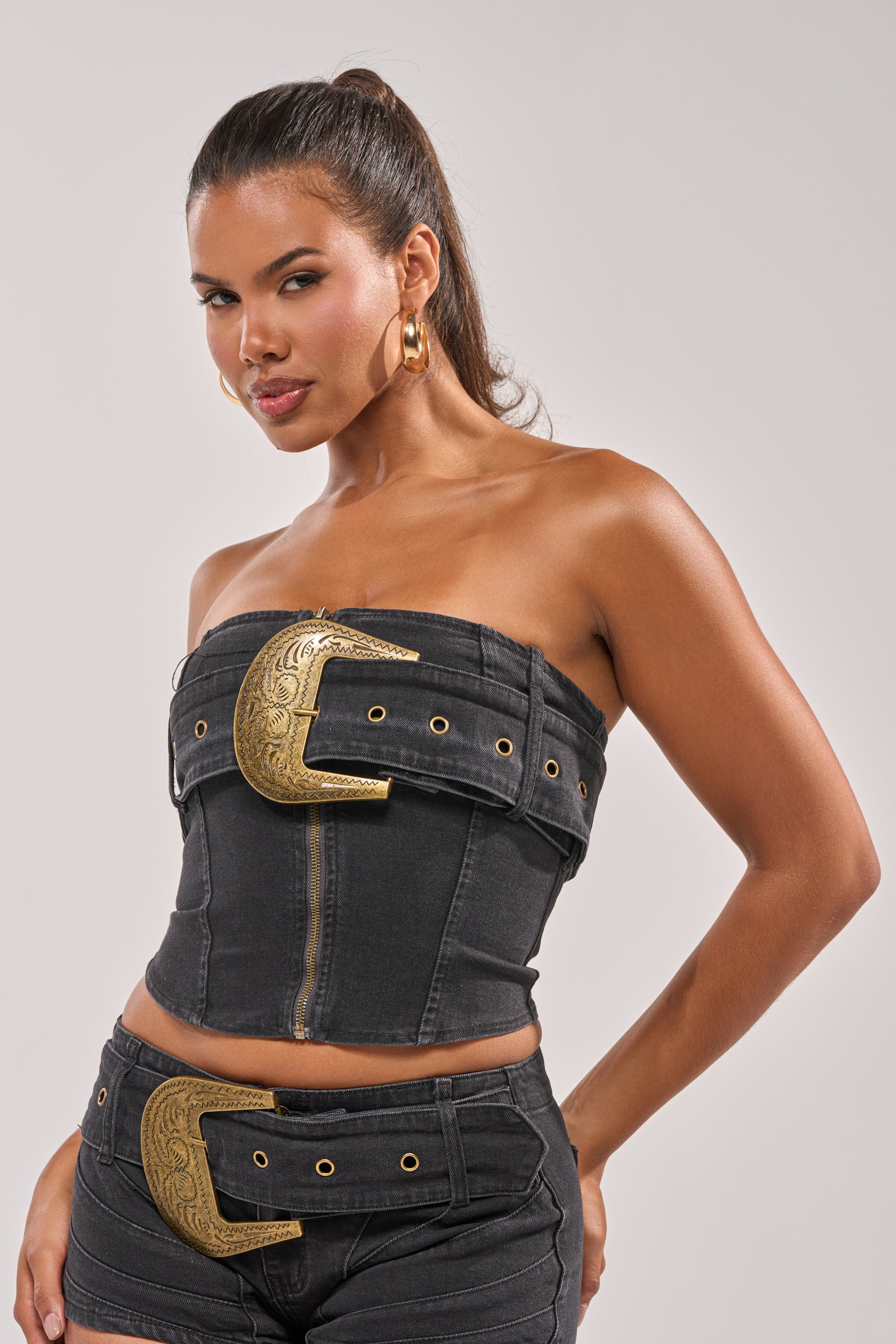 A woman with long brown hair in a ponytail poses confidently against a neutral background, wearing the BUCKIN DENIM TUBE TOP, a western-inspired black denim corset adorned with large, ornate gold belt buckles. 1