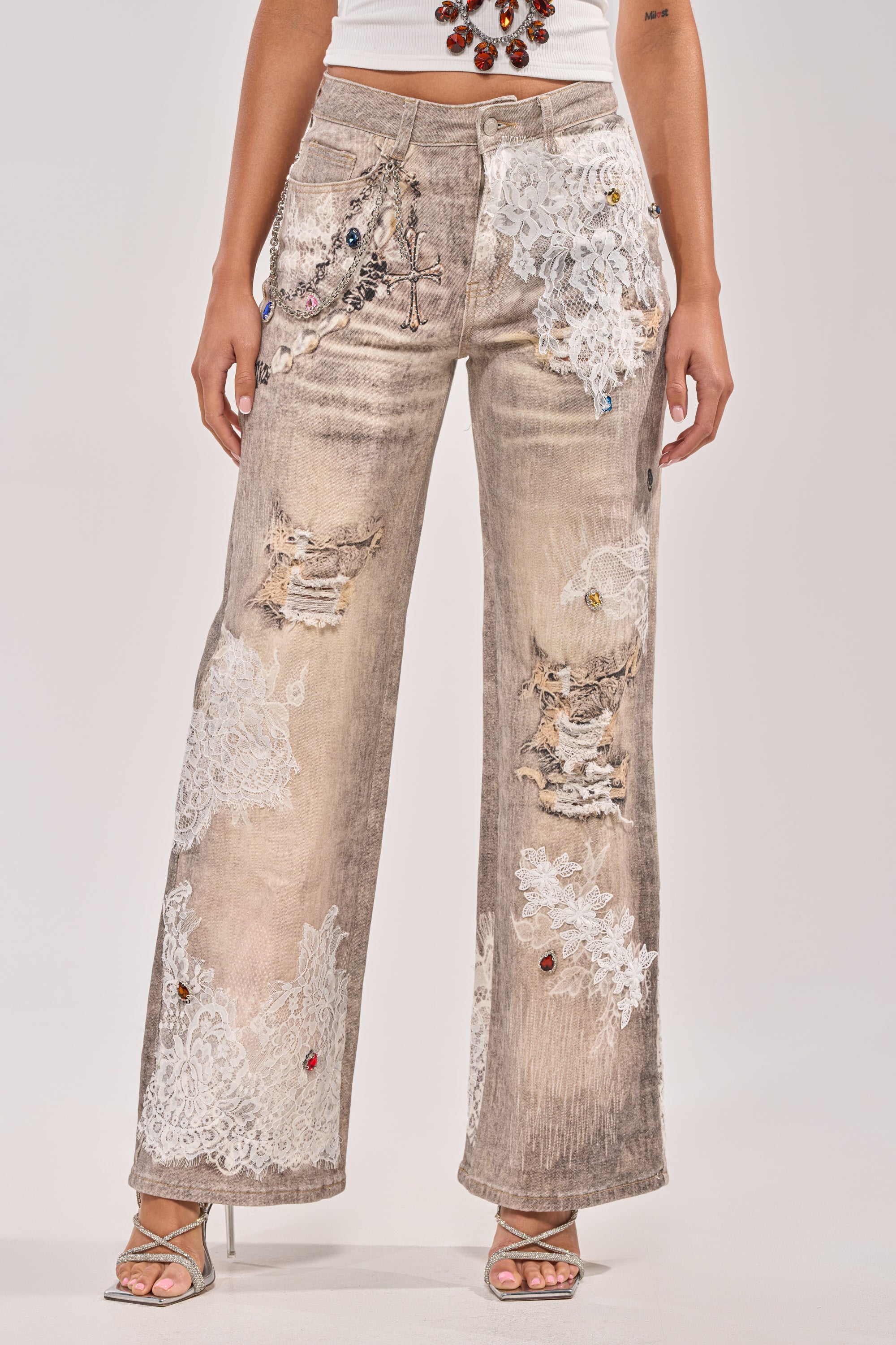 A person wears DONT MESS WITH ME RELAXED DENIM—wide-leg, distressed beige jeans with white lace, jewels, and embroidery—styled with strappy high-heeled sandals and a white top. The photo is cropped from shoulders to feet. 0