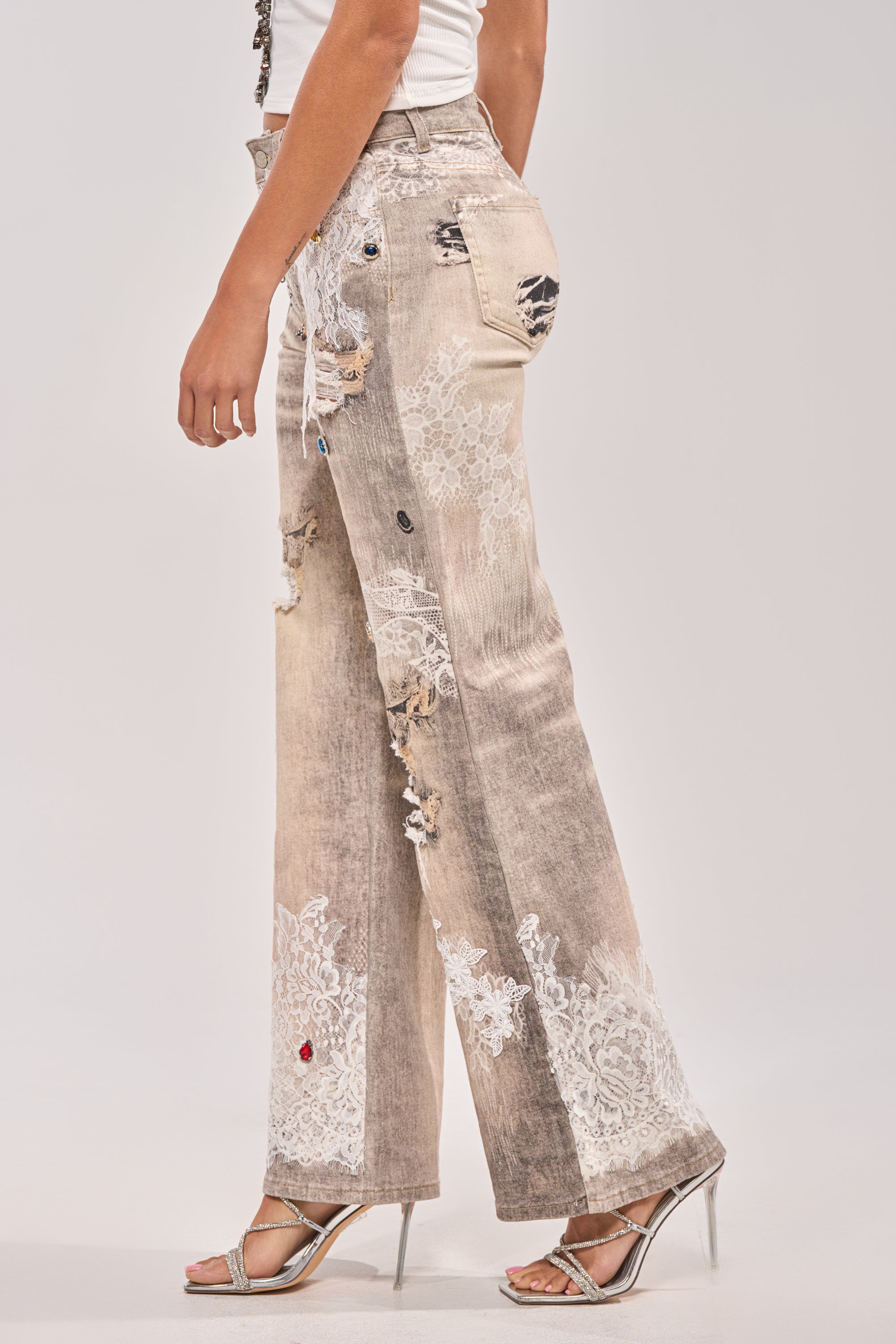 A person wearing the DONT MESS WITH ME RELAXED DENIM—high-waisted, lace-embellished distressed light brown jeans with white lace details—paired with silver high-heeled sandals, standing against a plain light background. 2