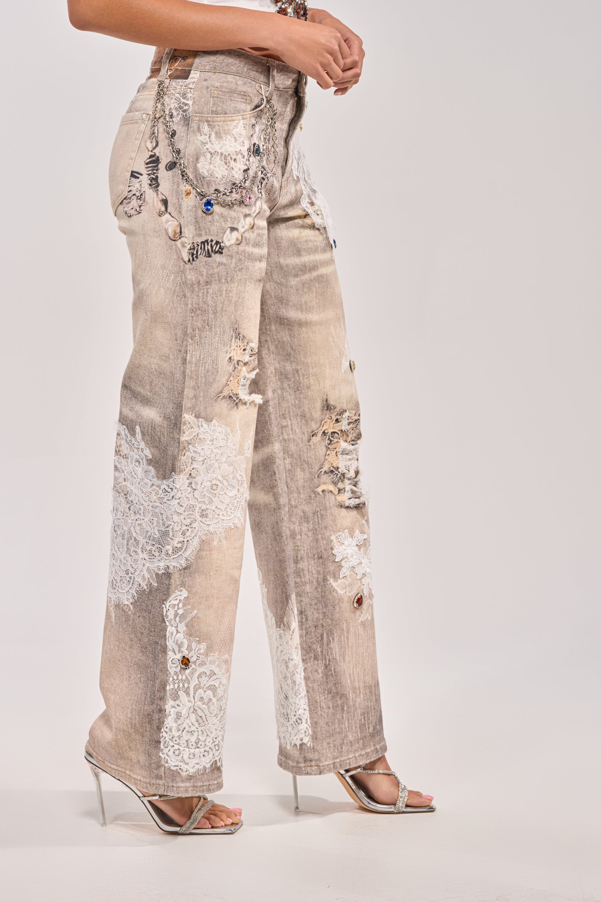 A person wearing DONT MESS WITH ME RELAXED DENIM jeans with lace embellishments and white lace patches, paired with silver high-heeled sandals, stands against a plain light background. 3