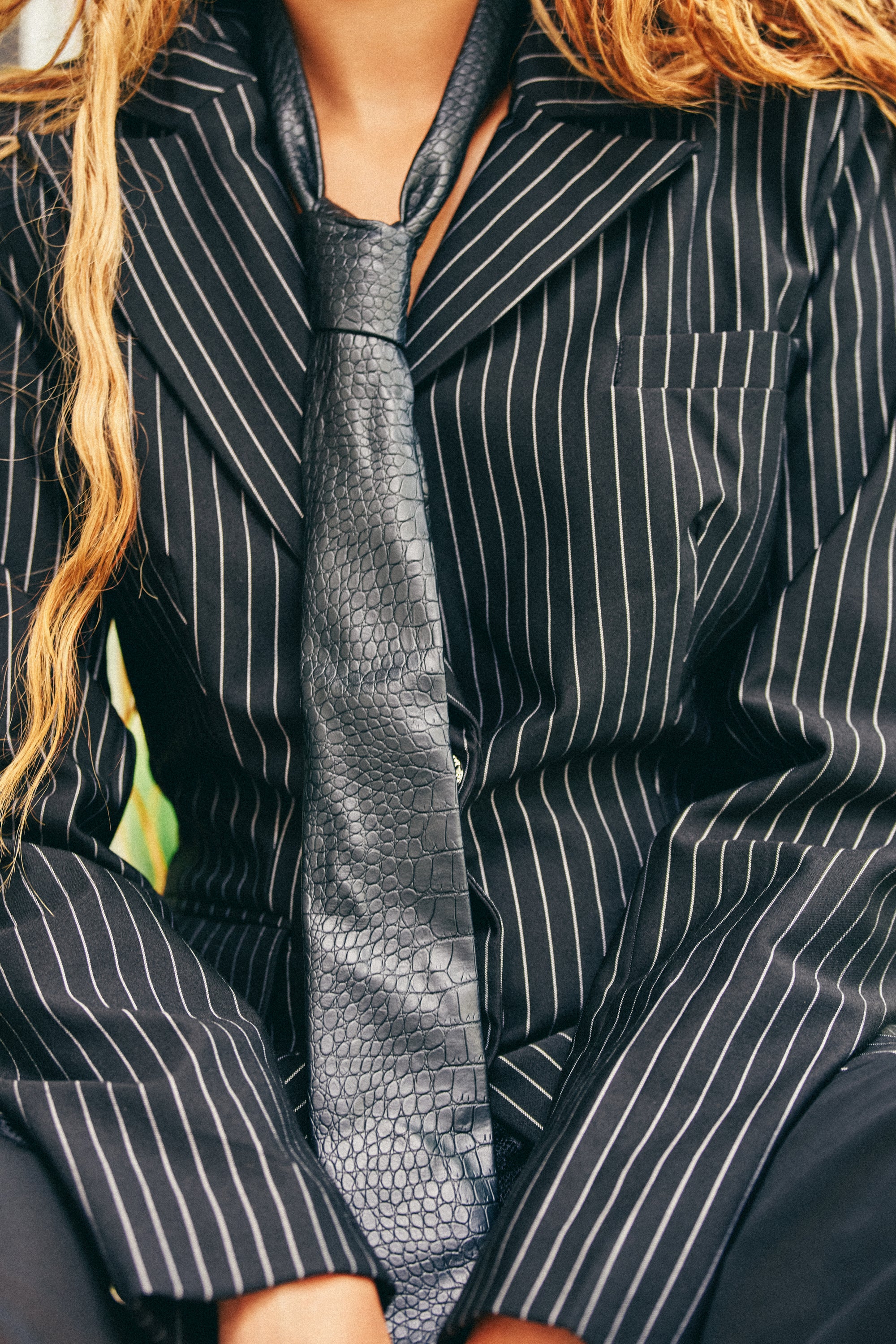 Wearing the ALIEN PU TIE—a sleek black snakeskin accessory—a person in a black pinstripe suit sits with relaxed arms. Their long, wavy hair is visible, and the photo is cropped to show only the upper body, not the face.