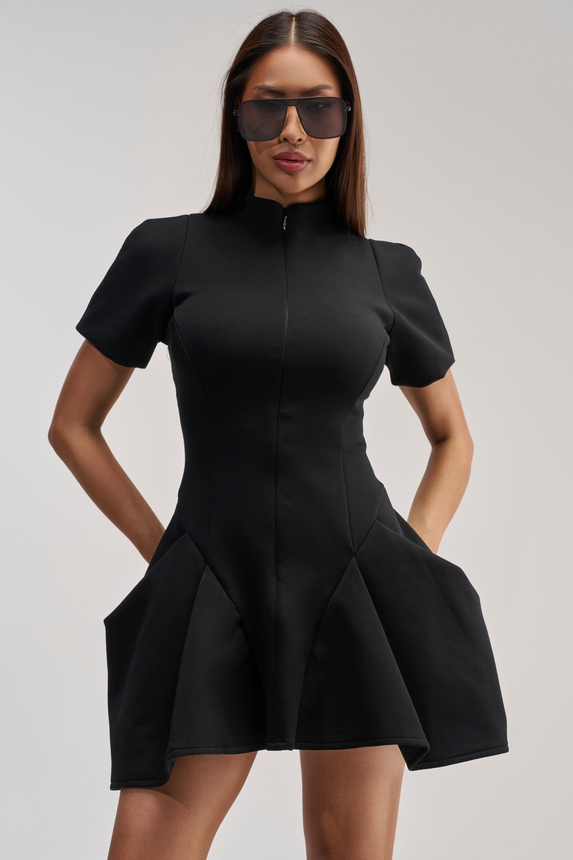 Model wearing the THAT WAS INTENTIONAL STRUCTURED MINI DRESS IN BLACK— a fit-and-flare silhouette featuring short sleeves, a zip-up front, flared skirt, and roomy pockets—poses with hands in pockets and oversized sunglasses against a plain background.