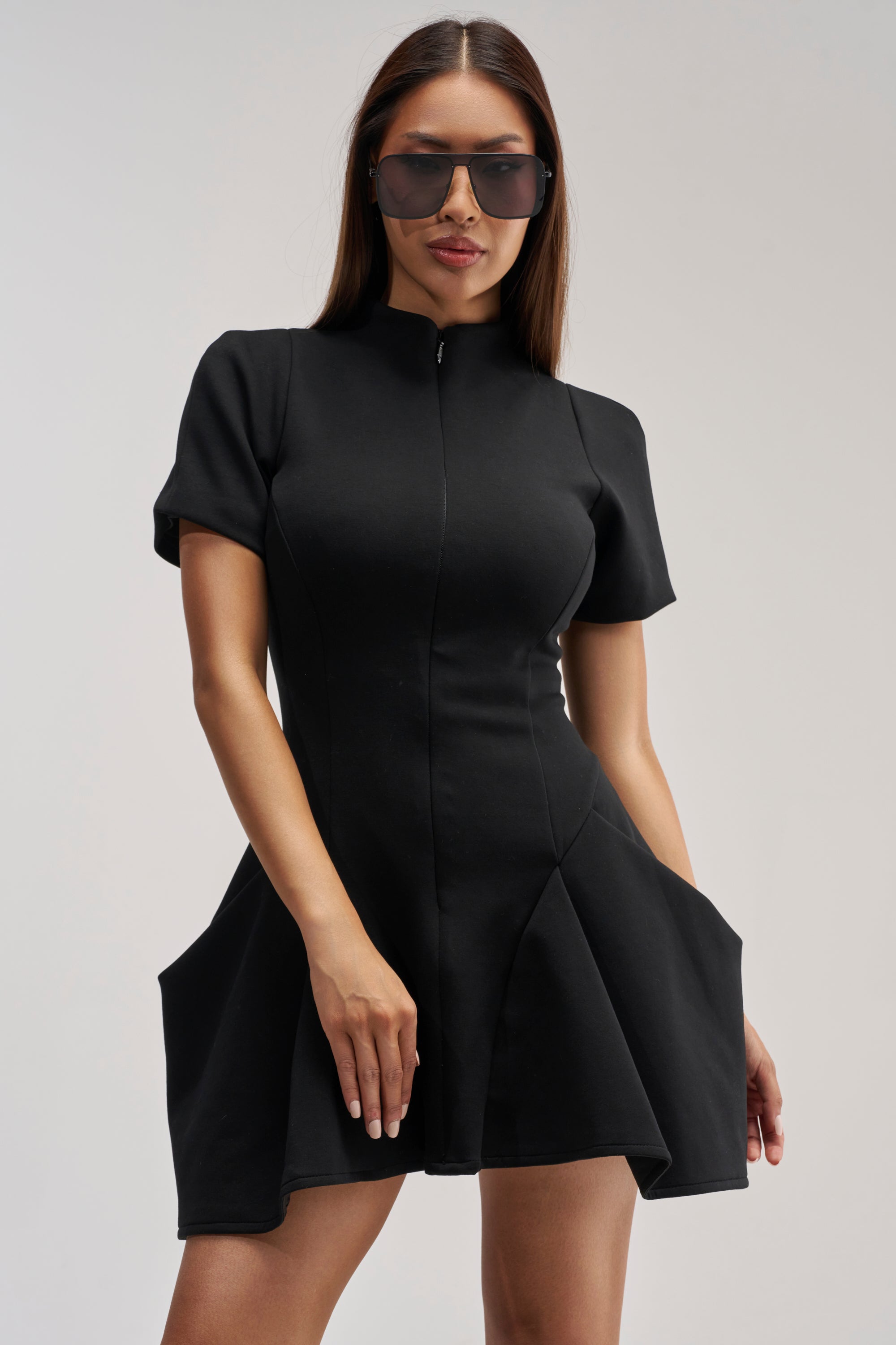 A woman wearing the THAT WAS INTENTIONAL STRUCTURED MINI DRESS IN BLACK—a short-sleeved, black fit and flare dress featuring a zippered front and a flared skirt—paired with sunglasses, stands against a plain light background.