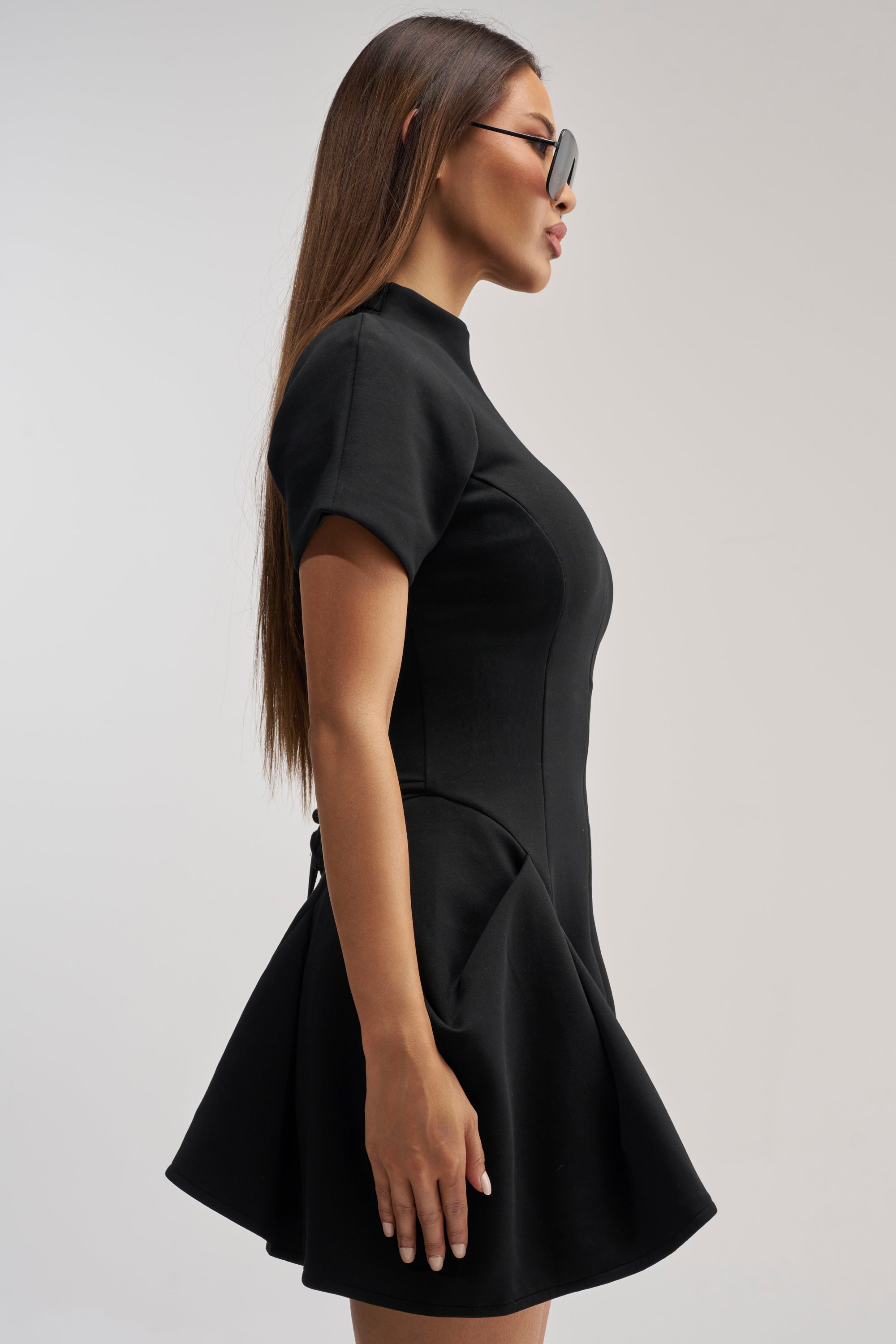 A woman stands in profile against a plain background, wearing the THAT WAS INTENTIONAL STRUCTURED MINI DRESS IN BLACK—a fitted, short-sleeve fit and flare style with a flared skirt—paired with sunglasses and her long straight hair worn down.