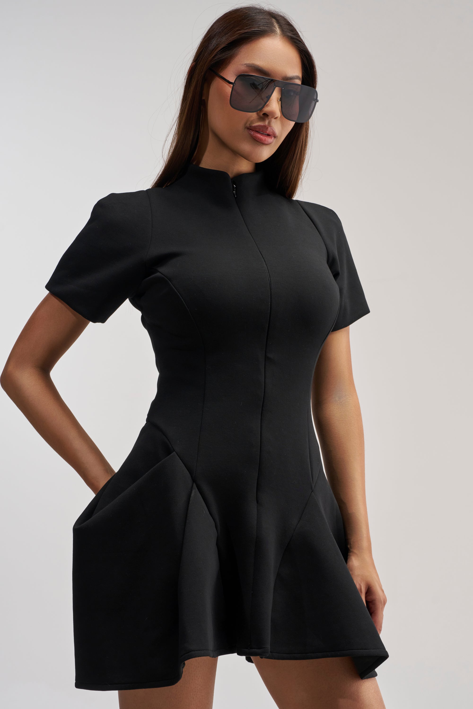 A woman models the THAT WAS INTENTIONAL STRUCTURED MINI DRESS IN BLACK—a fitted, short-sleeve cocktail dress with a flared skirt—posing confidently with one hand on her hip. She has long brown hair, wears oversized sunglasses, and stands against a plain light background.