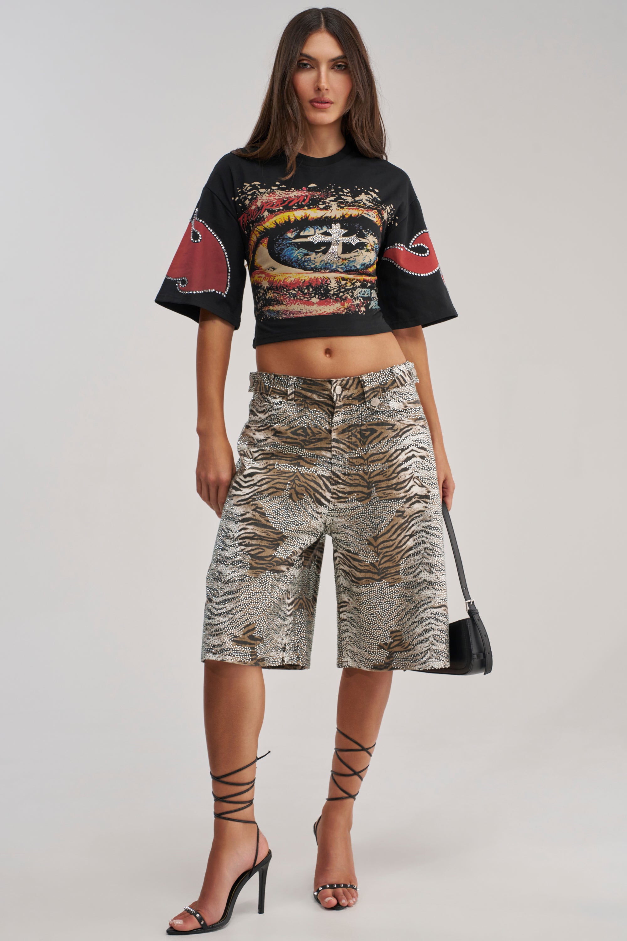 A woman poses against a plain background wearing a black graphic crop top, FEELING WILD RHINESTONE ZEBRA BERMUDA SHORTS, strappy high-heeled sandals, and carrying a small black handbag—perfect for festival streetwear.