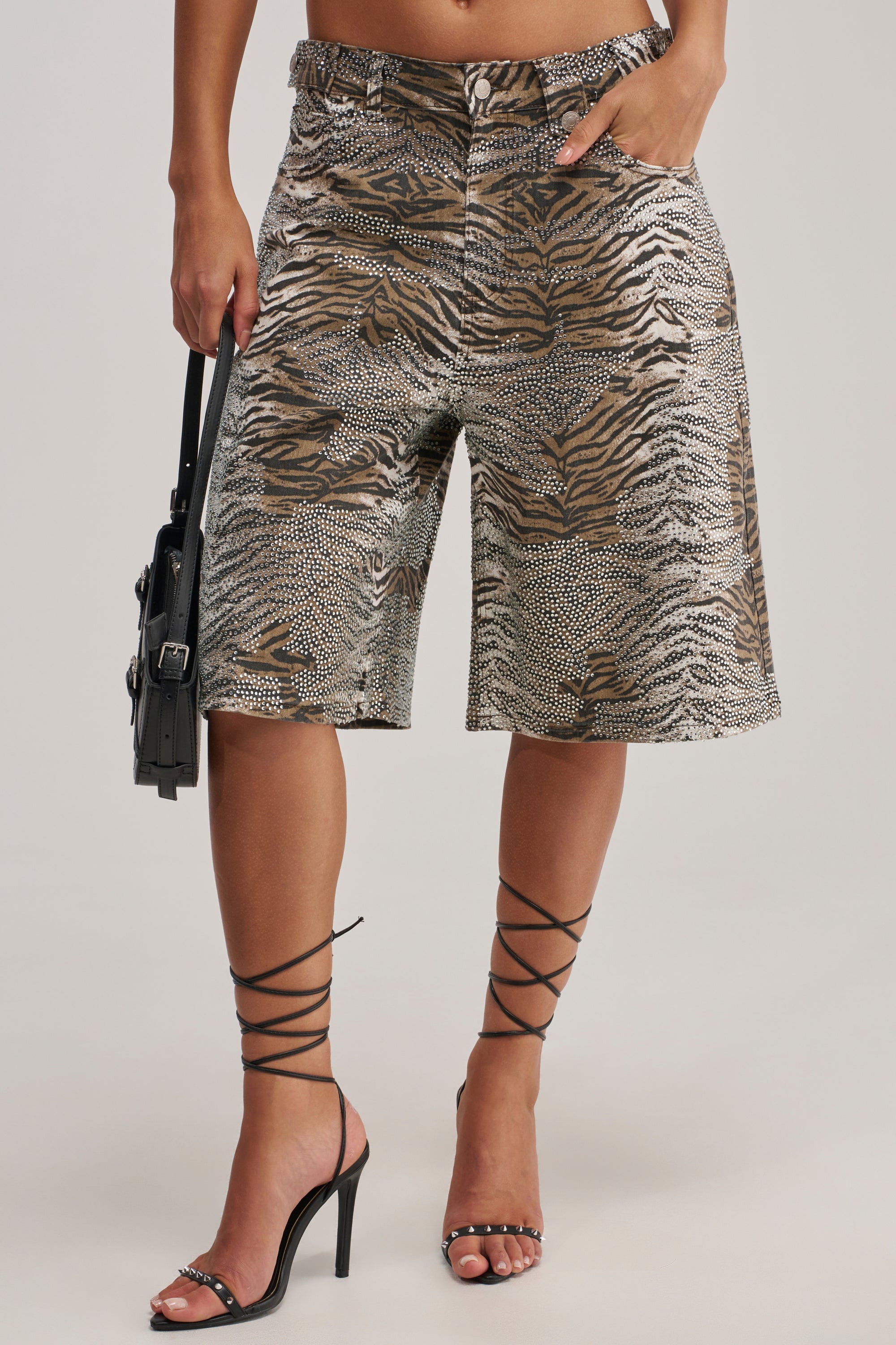 A person wears the FEELING WILD RHINESTONE ZEBRA BERMUDA SHORT styled as festival streetwear, paired with strappy black high heels and a small black handbag.