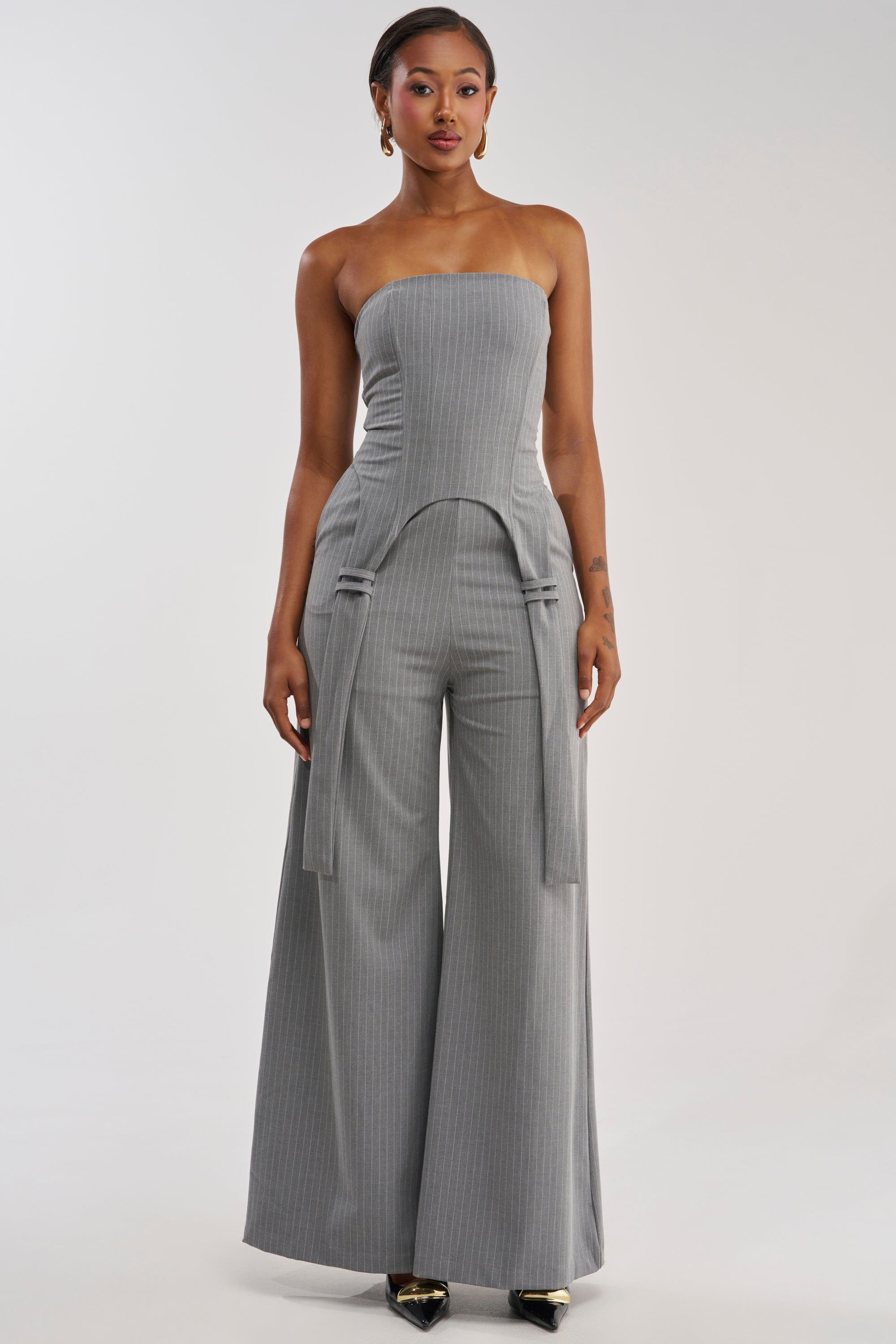 A woman models the WATCH AND LEARN STRAPLESS PINSTRIPE JUMPSUIT, featuring a strapless gray pinstripe corset top, wide-leg pants, and decorative hip ties, standing against a simple light background—the ideal outfit for a night out.